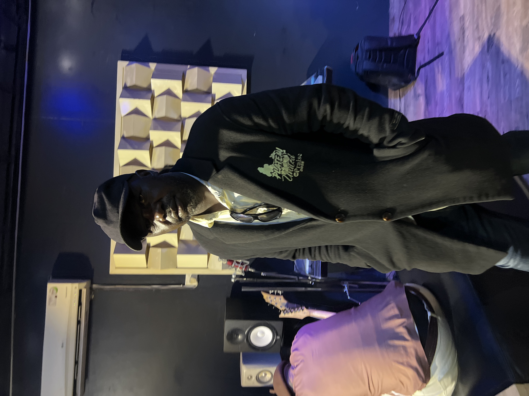 A man wearing a black baseball cap and a black overcoat over a yellow shirt stands in the center of a dimly lit room that appears to be a music studio. He has sunglasses tucked into his shirt pocket. The room has dark walls, a patterned acoustic treatment on the back wall, an air conditioner unit mounted high on the wall, and what appear to be studio monitors and musical instruments on the left side of the frame. A large, plush, reddish-brown object is positioned on the left, partially obscuring the studio equipment. The floor has a wooden texture. The overall impression is that of an indoor recording or rehearsal space. This scene is located in Jos, Nigeria.