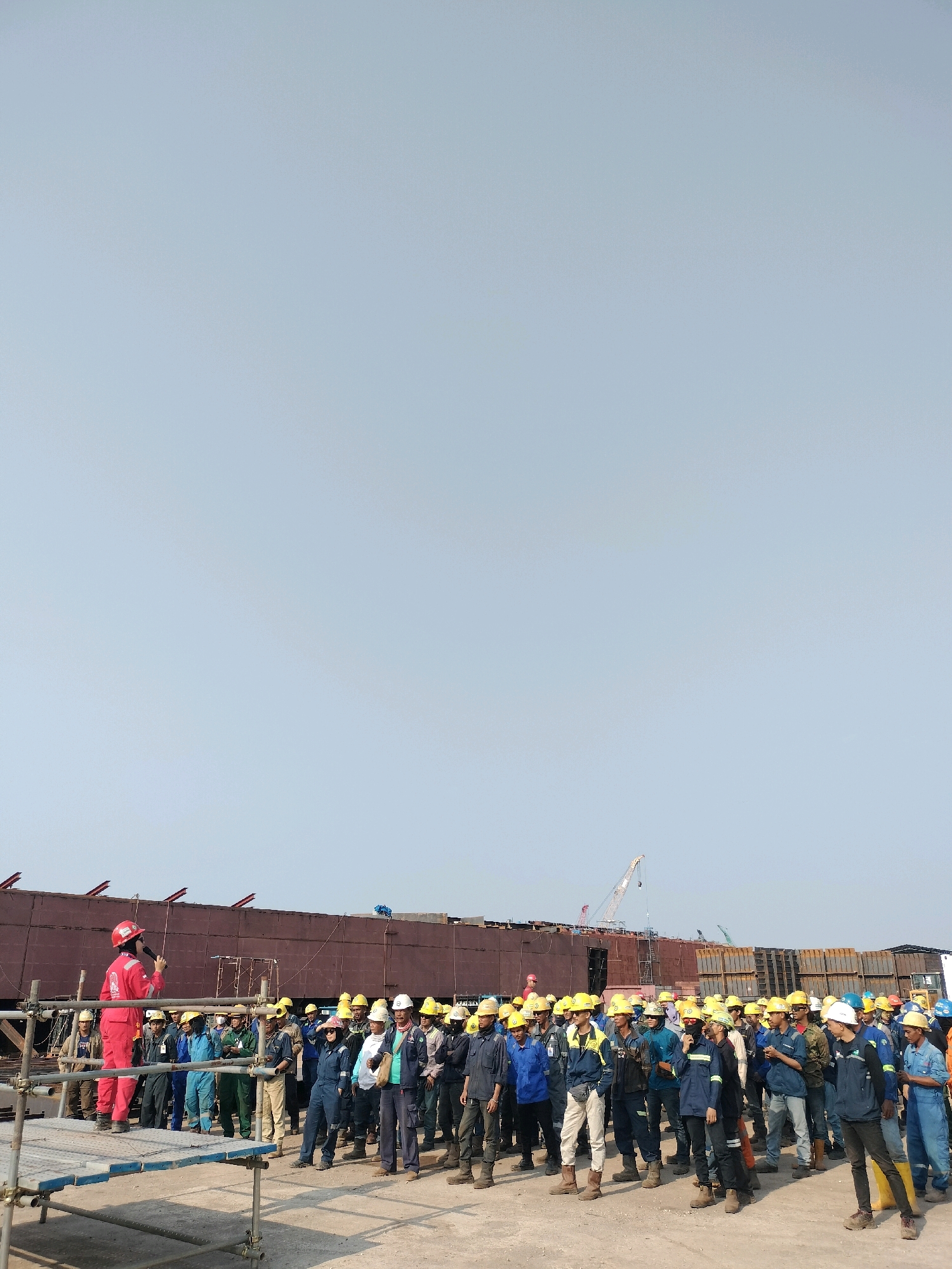 The image depicts a group of shipyard workers gathered outdoors. A woman in a red uniform and hard hat stands on a raised platform, addressing the group and holding a microphone, suggesting she is giving instructions or a briefing. The workers, most of whom are also wearing hard hats and safety vests, are standing and listening. In the background, a large ship under construction dominates the scene, with scaffolding and cranes visible, indicating an active shipbuilding or repair facility. The environment appears to be a shipyard, characterized by industrial structures and equipment. The city of Batam, Indonesia, is a well-known hub for shipbuilding and maritime industries, making a shipyard a plausible location for this scene. The weather appears to be clear and sunny.