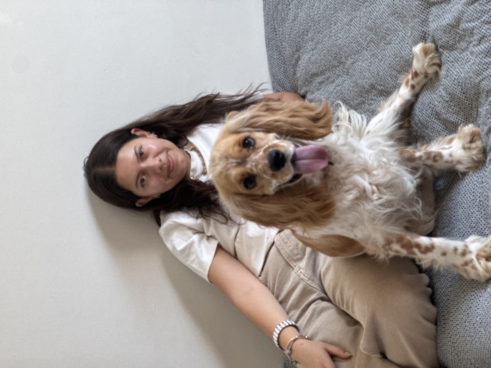 An indoor, casual portrait features a young woman and a dog. The woman, with long dark brown hair, is positioned on the left side of the frame, lying against a plain light-colored wall. She wears a white top, light brown or beige pants, a gold-toned necklace, small earrings, and multiple bracelets on her left wrist, including one with silver and black stripes. She is looking towards the camera with a gentle smile.
Next to her, on a grey textured upholstered surface (likely a sofa or bed), is a fluffy dog, resembling a Cocker Spaniel, with light brown and white fur, and darker brown spots. The dog is looking directly at the camera with its tongue out, indicating a happy or playful demeanor. Its body is mostly white with light brown markings, particularly on its ears, face, and legs.
The scene conveys a relaxed and affectionate moment between the woman and her pet. The lighting suggests daytime. The overall mood is one of contentment and companionship. There are no visible location cues within the image itself to confirm the provided context of Heroica Puebla de Zaragoza, Mexico, nor is there any visible text.