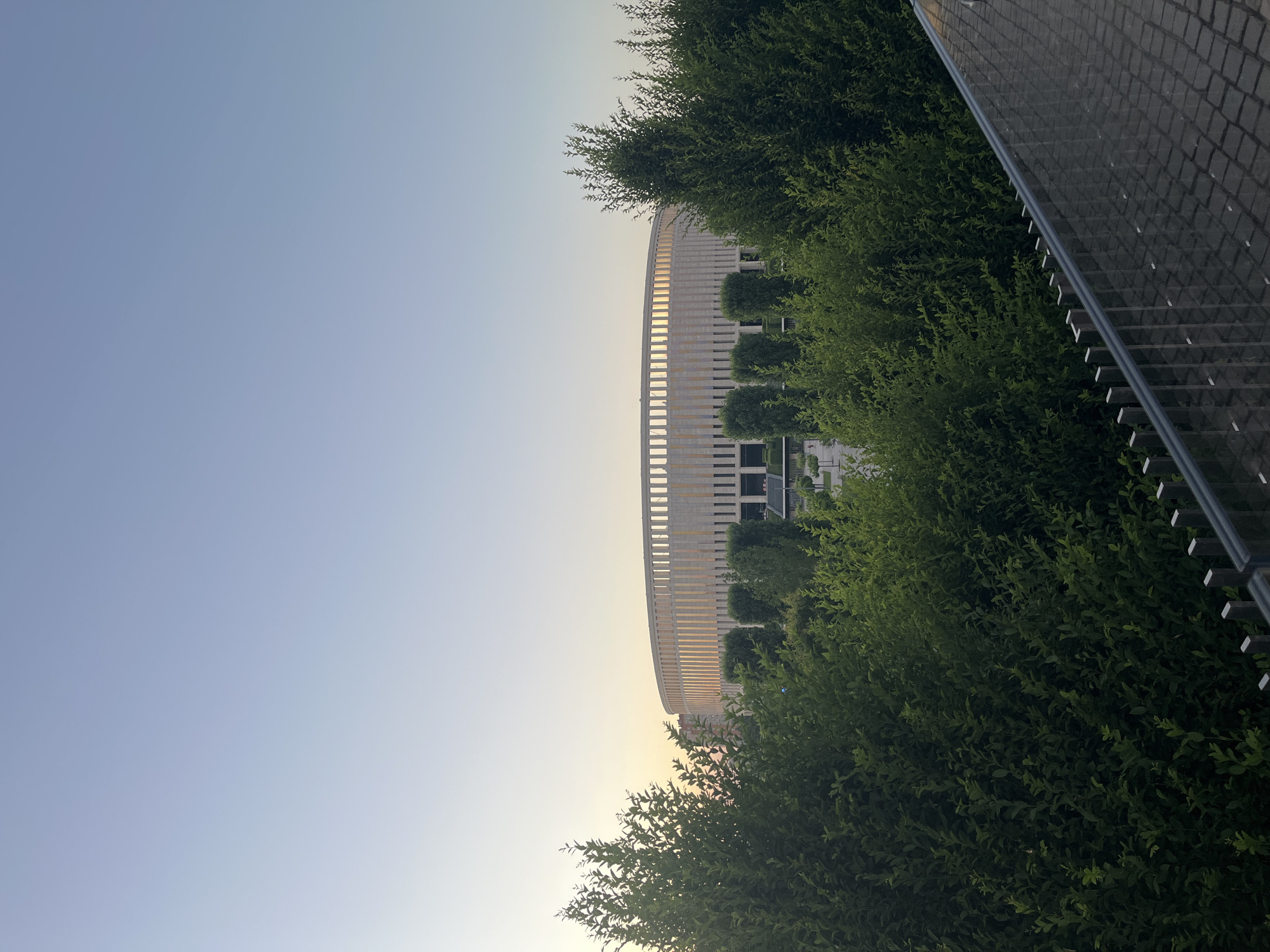 This image shows a wide shot of a modern stadium in Krasnodar, Russia, at dusk. The stadium is a large, circular structure with a light-colored facade and a row of vertical columns around its perimeter. The top of the stadium is illuminated by the setting sun, casting a warm glow on the building. In the foreground, lush green trees and bushes obscure the lower portion of the stadium, creating a natural barrier. A paved walkway with a metal railing is visible in the bottom right corner, suggesting it is a public area. The sky is clear and a pale blue, transitioning to a soft orange and yellow near the horizon, indicating the time of day. There are no people visible in the image. The scene is serene and focuses on the architecture and the surrounding greenery.