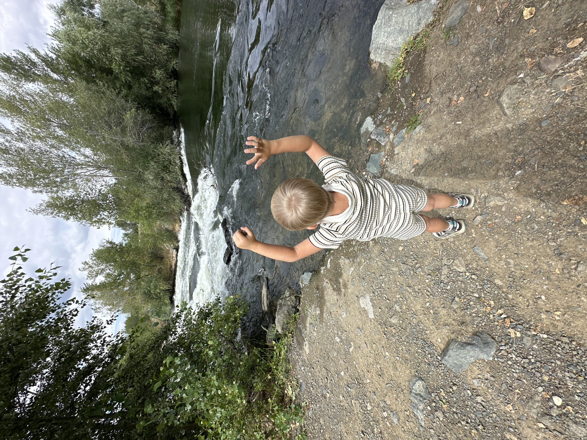 A young child, seen from behind, stands on a rocky shoreline with their arms raised. The child is wearing a striped onesie and sneakers. They are facing a fast-flowing river with rapids. Trees line the opposite bank of the river. The sky is partly cloudy. The scene appears to be a natural outdoor environment, possibly a park or recreational area within the city of Energetik, Russia.