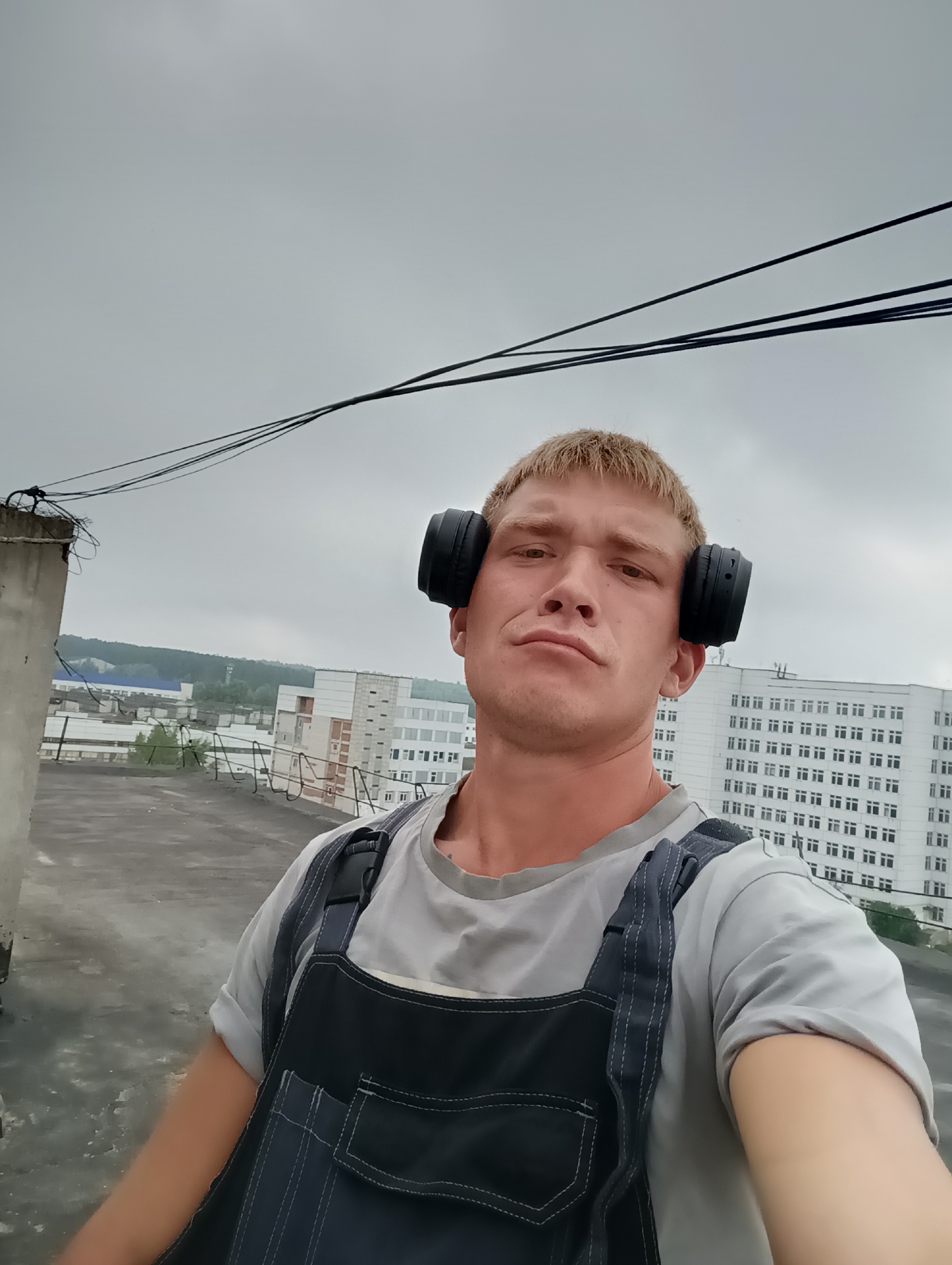 A fair-skinned male with short, blonde hair is captured from the chest up, wearing a gray t-shirt and dark blue overalls. He has black headphones on, covering his ears. The man is looking directly at the camera with a neutral expression. Behind him, a cloudy sky looms over a cityscape with several buildings, including a large white building with many windows. The man appears to be on a rooftop, with a concrete floor and railing visible in the foreground, and electrical wires stretching across the sky.