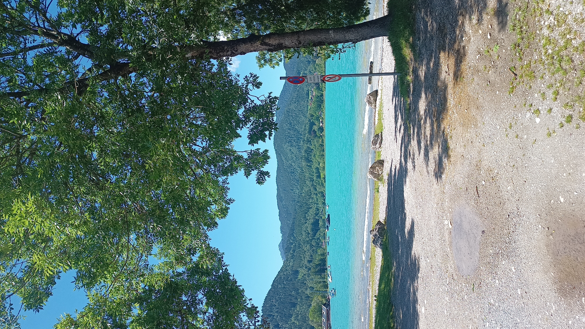 A picturesque view of the turquoise waters of Lake Tegernsee is captured from a tree-shaded bank in Rottach-Egern, Germany. The scene is framed by the lush green foliage of two large trees, one on the left and another on the right, with their branches creating a natural archway.
In the foreground, a gravelly area transitions into a grassy strip along the shoreline. Several large, light-colored rocks are scattered on the pebble beach. A signpost with two circular prohibition signs is visible on the right side of the image. The lake is calm with several sailboats anchored in the distance. Across the lake, a range of verdant, tree-covered hills and mountains forms the backdrop under a clear blue sky. On the far left, a building with a wooden facade is partially visible, situated at the water's edge. The lighting suggests it is a bright, sunny day. There are no people visible in the image.