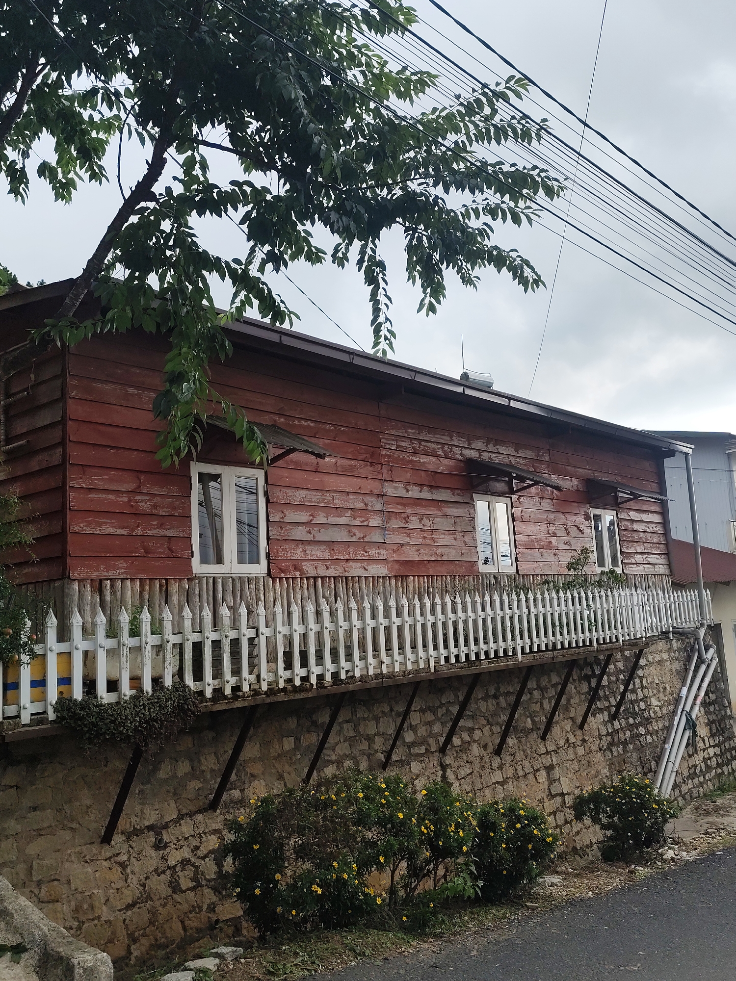 A two-story wooden house with red siding is built on a hillside overlooking a paved road in Xuân Thành, Vietnam. The house has a white picket fence along its front porch, which is supported by a stone wall. There are several trees and bushes growing around the house, and power lines run overhead. The sky is cloudy.
