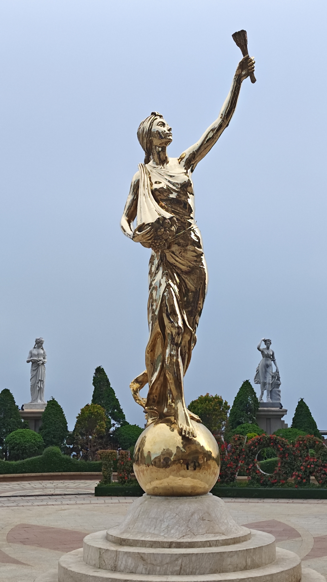 This image showcases a grand outdoor sculpture garden, likely located in Đà Nẵng, Vietnam, dominated by a large, golden statue against an overcast sky.
The central subject is a prominent golden statue of a woman, rendered in a classical style. She stands dynamically on a large, reflective golden sphere, her bare feet visible. Her right arm is extended upwards, holding a torch-like object, while her left arm is bent, cradling a fold of her draped garment which appears to contain grapes or fruit. Her head is tilted upwards, and her gaze is directed towards the sky, conveying a sense of inspiration or dedication. The statue's highly polished surface reflects the surrounding light and environment.
The golden sphere rests on a multi-tiered circular base made of light-colored marble. The ground immediately around the base is a paved area with a decorative pattern of reddish-brown and lighter-toned tiles.
In the mid-ground, behind the central statue, a meticulously landscaped garden is visible. It features manicured green hedges, tall, conical evergreen trees, and vibrant red floral arrangements, including a circular one that resembles a wreath. Two smaller, lighter-colored statues of women, also in classical attire and posed, stand on pedestals in the distance on either side of the main figure.
The sky is a uniform, pale blue-gray, indicating an overcast day with soft, diffused lighting. The scene appears serene and well-maintained, suggesting a public park, botanical garden, or a resort complex. No text is visible in the image.