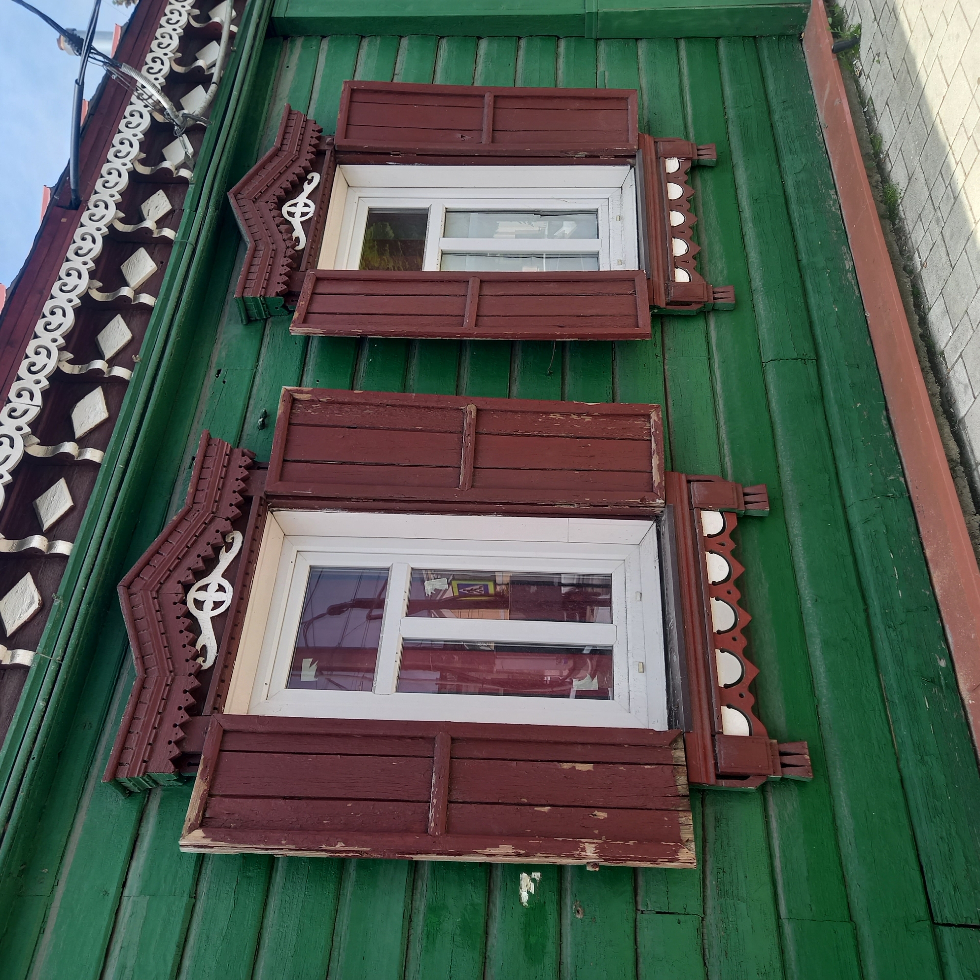 This image depicts a detailed, angled view of the exterior of a traditional wooden building in Zvenigorod, Russia, under clear daylight conditions. The building's facade is constructed from horizontal wooden planks, painted a weathered, dark green with visible chipping and fading.
Two rectangular windows with modern white PVC frames and clear glass panes are prominent. Both windows are flanked by open, dark reddish-brown wooden shutters, which also show significant paint loss and weathering, revealing the natural wood underneath in places.
Each window is adorned with elaborately carved wooden decorative elements, or platbands. Above each window, the platbands are painted dark reddish-brown with white accents, featuring a pointed, gable-like top section with intricate scrollwork and a central four-lobed motif. Below each window, the platbands are similarly carved and colored, with a pattern of semicircular cutouts and vertical supports extending downwards from the sides.
Above the green wall and the upper window decor, a dark reddish-brown fascia board or eaves section is visible, featuring a repeating pattern of white carved diamonds and scrollwork. Electrical wires and cables are strung horizontally across this upper section, particularly above the right window, with some wires secured to the facade.
The glass of the left window distinctly reflects a blue square pedestrian crossing sign with white figures, indicating activity or structures opposite the building.
At the very bottom of the frame, a dark reddish-brown wooden plank or trim runs horizontally along the base of the green wall. Below this, a grey paved sidewalk, composed of rectangular blocks, is visible, with small patches of green vegetation growing in the cracks on the right side. The sky, partially visible at the top right, is light blue with sparse white clouds. No individuals are present in the scene.