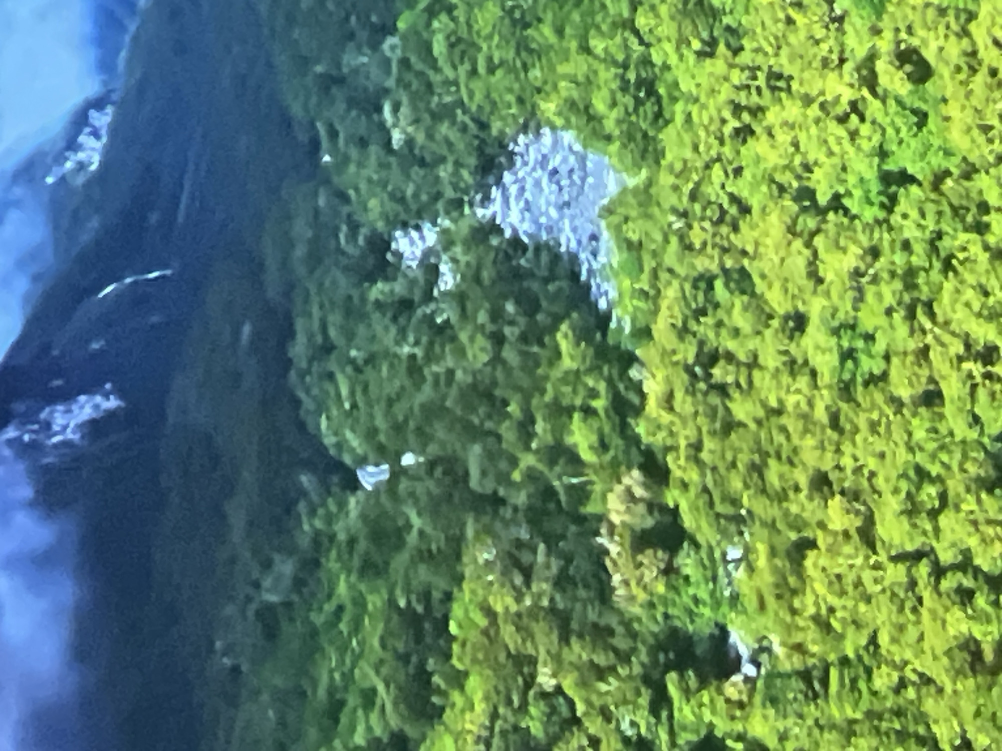 The image displays a mountainous landscape covered in dense green foliage, with a river flowing through it. The sky above the mountains is overcast and appears a hazy blue. There is no evidence of urban environments or specific landmarks that would indicate a location within Casablanca, Morocco.