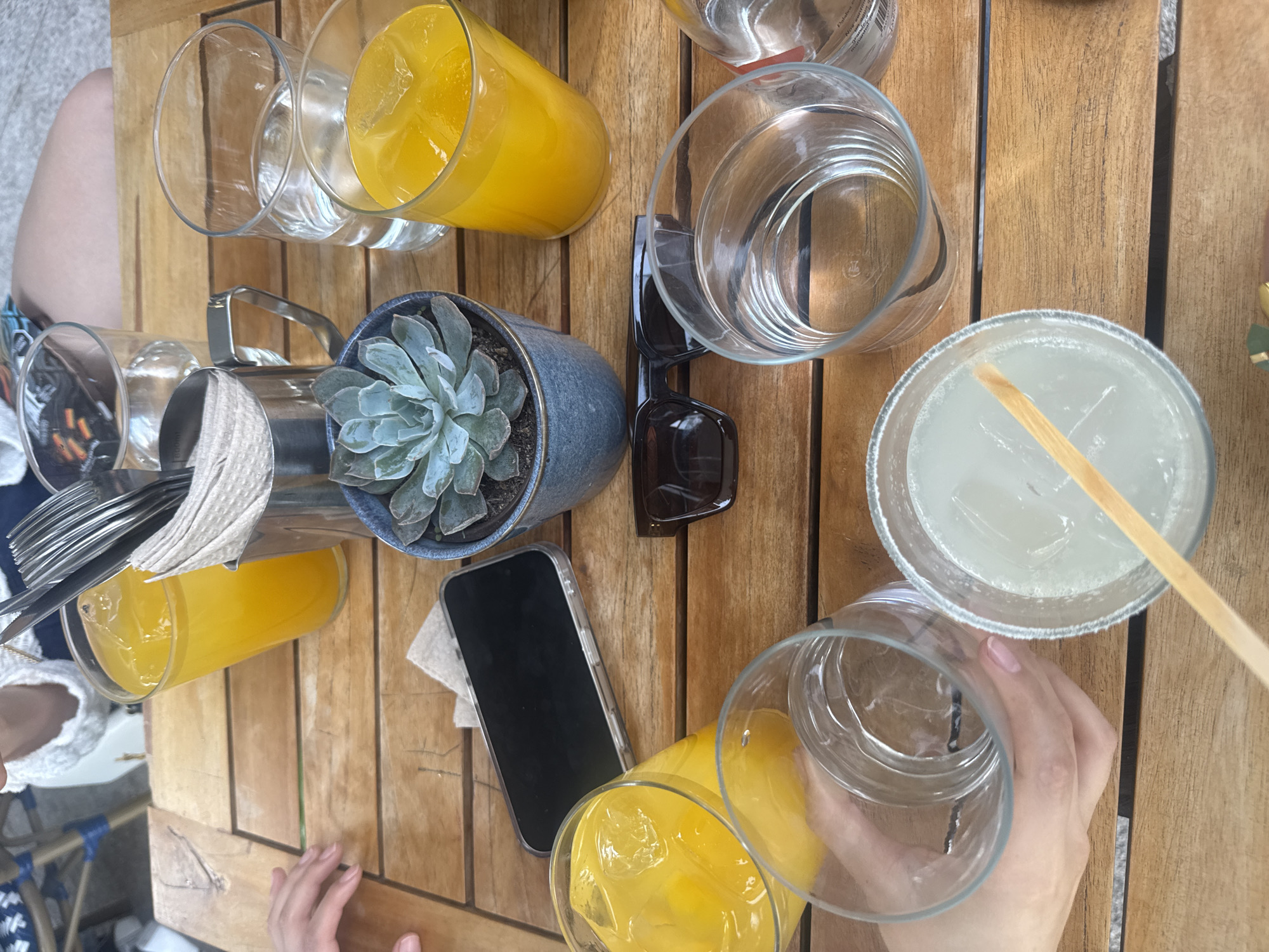 The image depicts a top-down view of a wooden table, presumably at an outdoor dining establishment, with various items scattered across it.
Subjects/Objects:
* Multiple glasses, some filled with orange juice and others empty or containing water.
* A potted succulent.
* A pair of sunglasses.
* A smartphone.
* A silverware caddy with silverware and napkins.
* A drink that appears to be a margarita.
* Two sets of hands that can be seen in the photograph that are reaching for drinks.
Actions/Interactions:
* People are reaching for drinks on the table.
Environment:
* The setting appears to be a casual outdoor dining area, possibly in Mexico City, Mexico. The weather seems to be nice, judging by the sunglasses, open seating, and the light.