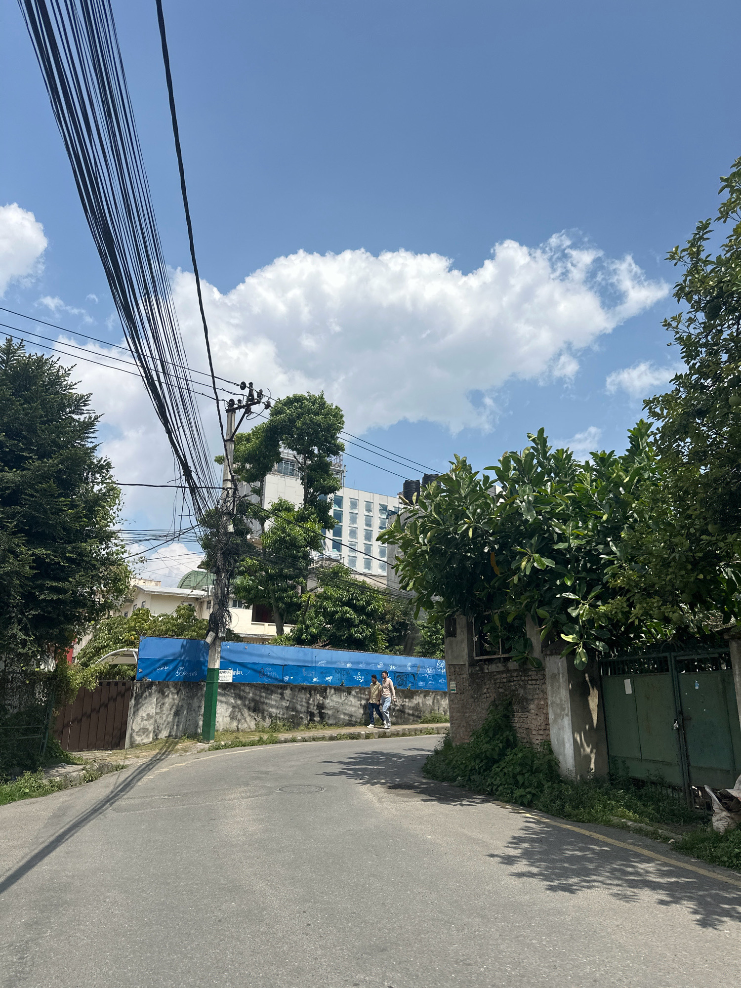 The image shows a street scene in Lalitpur, Nepal. A paved road curves into the distance, with a line of trees on the right and a wall with a blue banner on the left. Overhead, a dense network of power lines crisscrosses the sky, casting shadows on the road. In the background, a modern white building with many windows rises above the trees. Two individuals are walking on the sidewalk near the blue banner. The sky is blue with fluffy white clouds.