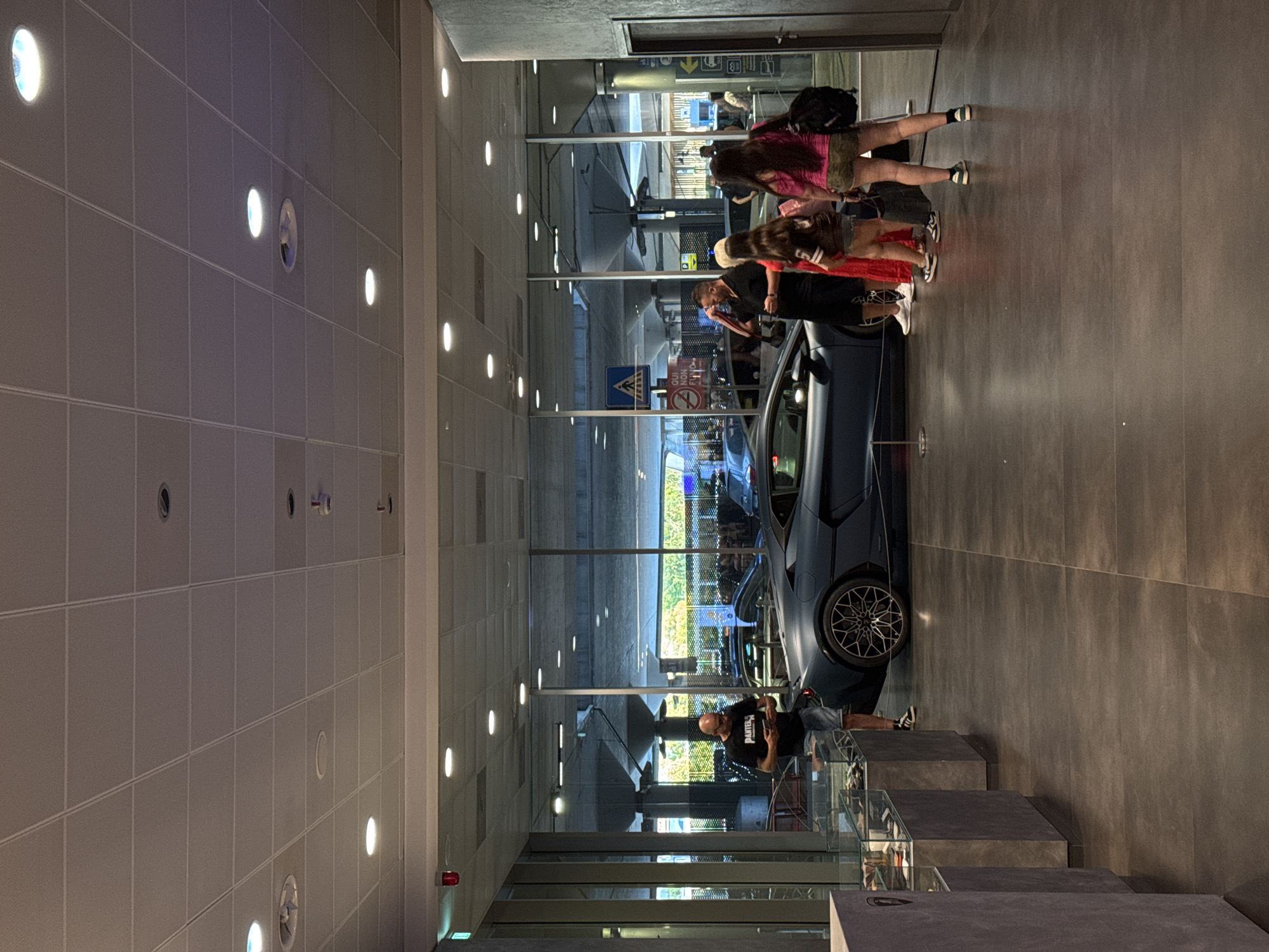 The image depicts a modern interior with large glass windows looking out onto an exterior area. Inside, a dark blue Lamborghini is parked, with several people standing around it. To the left, a man in a black t-shirt and shorts is looking at his phone. Further to the right, a group of three women are also gathered near the car. The floor is tiled, and the ceiling features recessed lighting and what appear to be air vents. The overall impression is that of a car showroom or a similar commercial establishment. The location is identified as Bologna, Italy, suggesting this is a car dealership or exhibition space within the city.