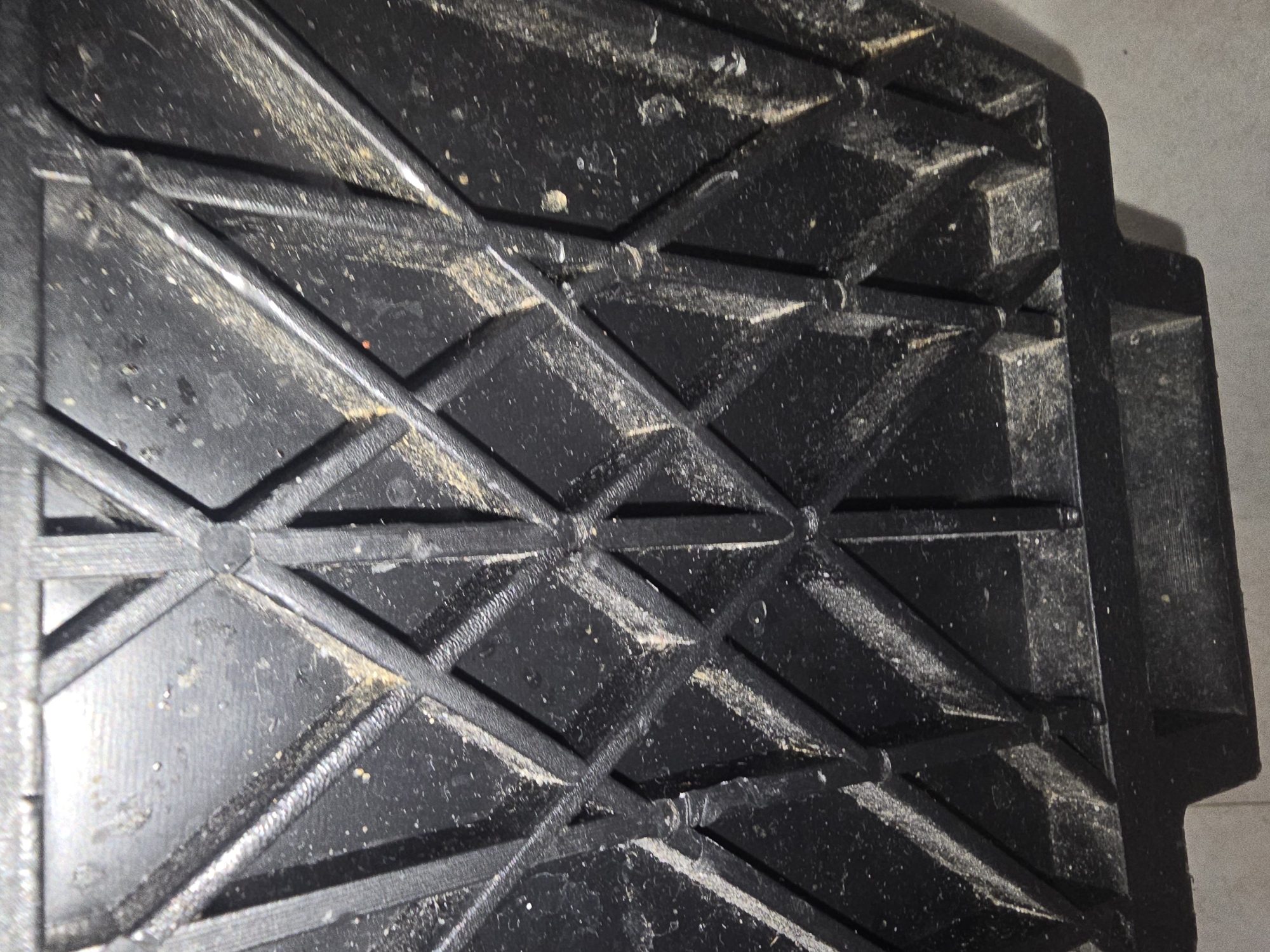 This is a close-up, overhead view of the underside of a black plastic object, likely a storage container or a piece of equipment. The surface is characterized by a raised, diamond-shaped grid pattern, with intersecting raised bars. The plastic is matte and appears to be somewhat worn, with visible scuff marks and dust accumulation, particularly along the edges and within the recessed areas of the grid. The dust is a light brown or tan color. The object is positioned indoors, as suggested by the ambient lighting and the presence of what appears to be a tiled or concrete floor in the lower part of the image. There are no people or animals present in the image. There is no information to indicate the location in the city of Ulan-Ude, Russia.
