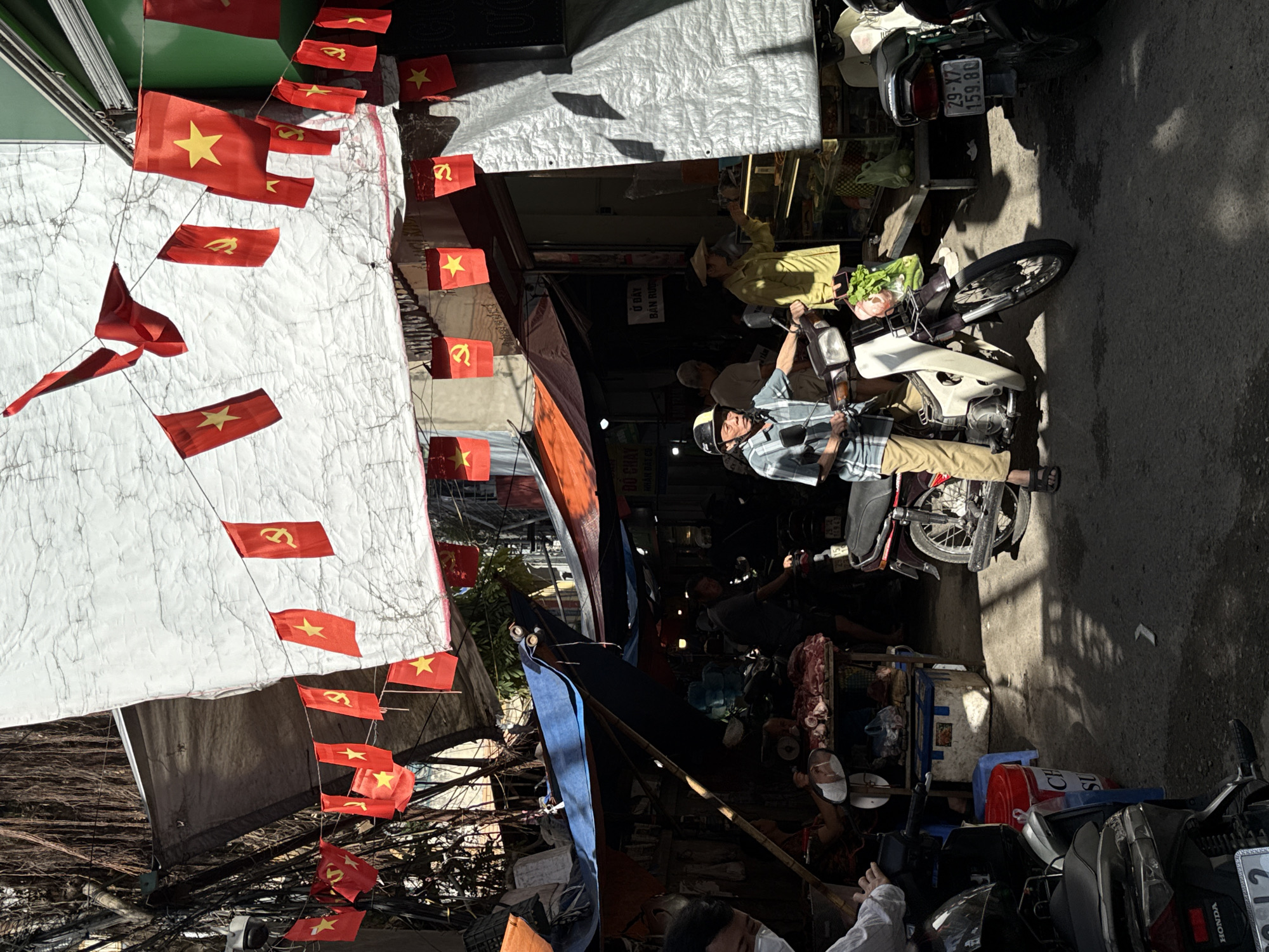 The image shows a street scene in Hà Nội, Vietnam. The scene is bustling with people and motorcycles parked along the narrow street. Banners of Vietnamese flags are strung across the top of the street. People are sitting on their motorbikes. A woman is wearing a non lá hat. In the foreground, there are parked motorbikes, and you can see signs like "HONDA" and partial Vietnamese text. A store's sign reads "Ở ĐÂY BÁN RƯỢU" (Here sells alcohol). The photograph captures the energetic atmosphere of a typical commercial area in Hà Nội.