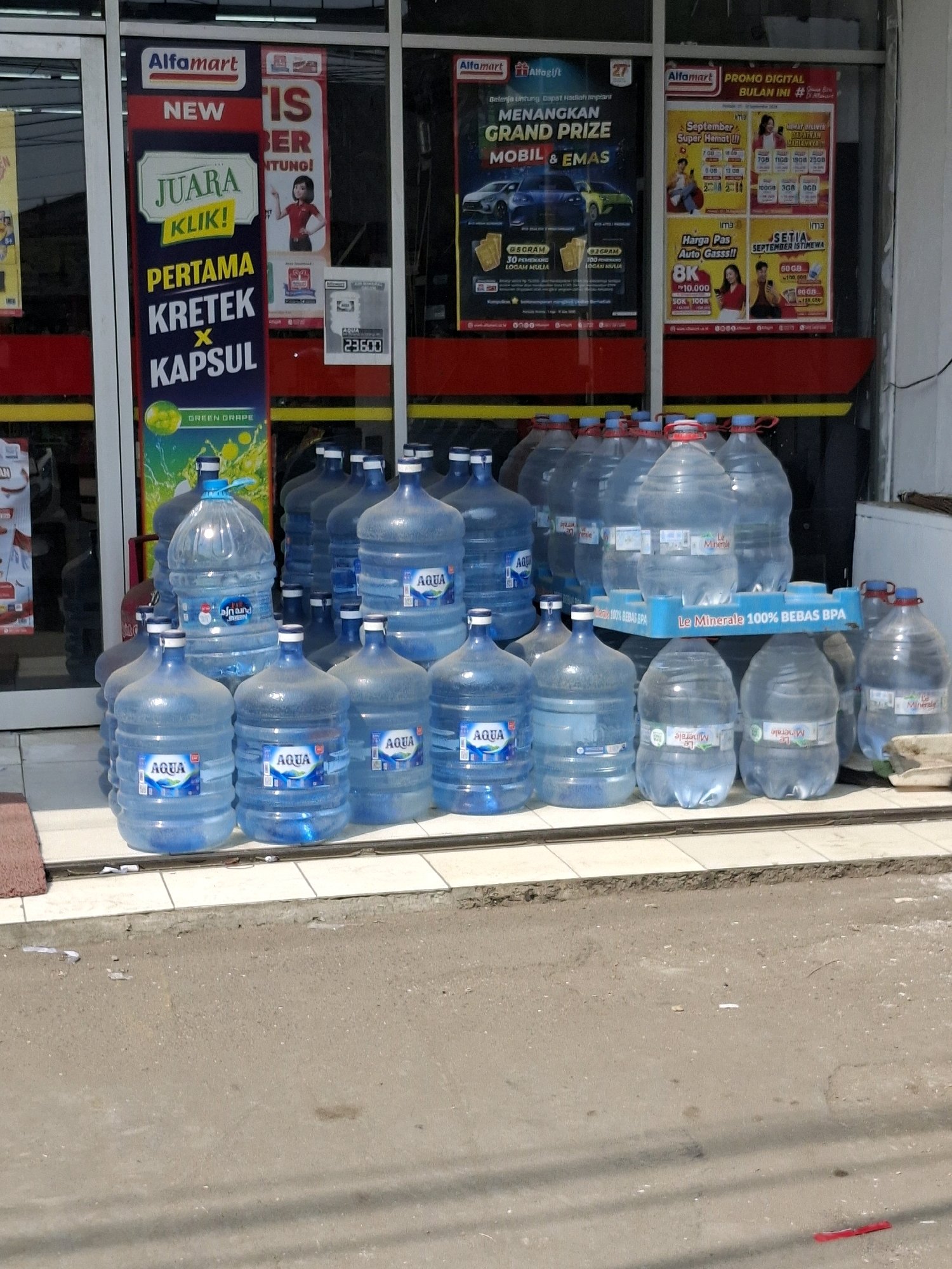 The image shows a storefront of an Alfamart convenience store in Selajambe, Indonesia. The store has a glass facade with promotional posters displayed on the interior windows. Outside the store, arranged in rows on the pavement, are numerous large, clear plastic bottles of drinking water. The bottles are of varying sizes, with some clearly labeled "AQUA" and others with "Le Minerale". The majority of the bottles are full and appear to be for sale. The ground in front of the store is a paved surface, possibly a sidewalk or street, with some debris scattered on it. In the background, through the glass, one can see aisles and shelving within the store. The lighting suggests it is daytime.