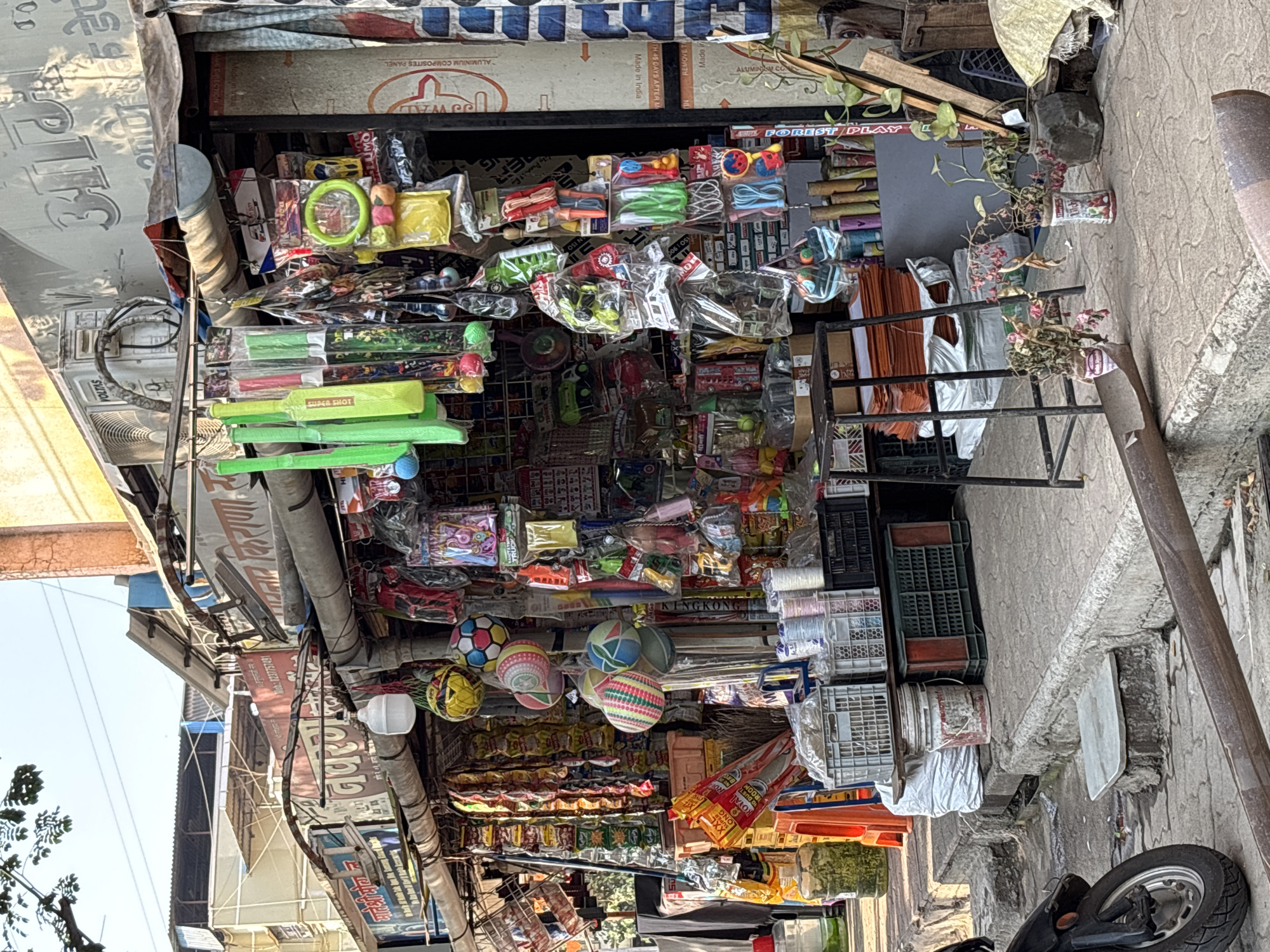 The image shows a roadside vendor stall in Ambernath, India, densely packed with various toys and goods. Visible items include plastic cricket bats, balls, toy cars, games, packaged snacks, and other miscellaneous merchandise. The stall is covered by a metal awning, and signage is visible, including brand names like Voltas and Royal. A motorcycle is parked on the street next to the stall. Crated plastic containers are near the shop. The street and building facades are visible, indicating an urban setting.