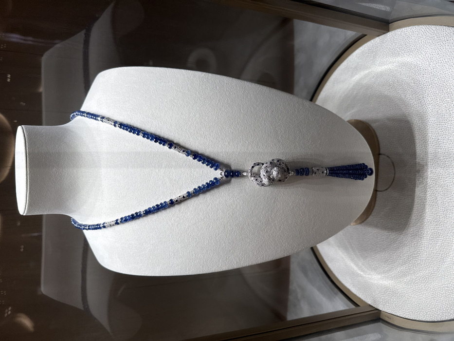 A high-jewelry necklace is displayed on an off-white, contoured jewelry bust within a glass display case. The necklace presents a long, Y-shaped design, primarily composed of numerous graduated, faceted dark blue sapphire beads. Sections of the necklace are accented with polished white metal elements pave-set with small, round brilliant-cut diamonds. The central pendant features an intertwined, stylized panther figure, entirely encrusted with pave-set diamonds and detailed with dark accents, likely onyx, for its spots and features. A multi-strand tassel of smaller dark blue sapphire beads descends from the bottom of the panther pendant. The bust rests on a subtly textured, light-colored circular base, which is enclosed by the display case's frame. Reflections on the glass indicate an upscale interior, consistent with a luxury retail environment. Given the distinctive "Panthère" motif, this piece is identifiable as a Cartier creation, placing its display within a Cartier boutique in Paris, France, likely in the vicinity of Place Vendôme. No individuals are visible in the scene.