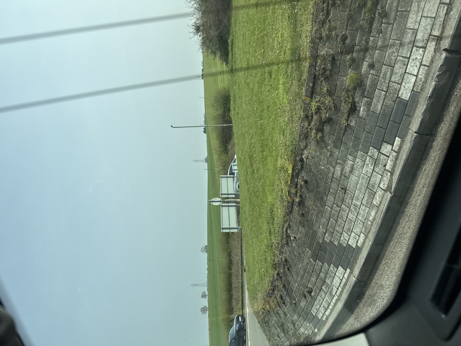 This image captures a wide-angle view from inside a vehicle at a roundabout on the outskirts of Thrapston, United Kingdom, likely within the A14/A45/A605 junction complex.
In the immediate foreground, a grey concrete retaining wall with a distinctive pattern of alternating light and dark rectangular blocks extends horizontally. Sparse grass and weeds grow along its top edge. A dark grey curb delineates the wall from a visible section of dark, damp-appearing asphalt road surface at the bottom of the frame. Vertical light streaks, consistent with reflections on a windshield, are visible in the upper right.
The midground features a large, green grassy island, characteristic of a roundabout's center. On the left side of the image, a dark-colored SUV, possibly black, is observed on the asphalt road, indicating movement from left to right around the roundabout. Positioned centrally on the grassy island are two white rectangular road signs mounted back-to-back on a single pole. Behind these signs and slightly to their right, a silver or light-colored sedan is partially visible, resting on a grassy incline with only its roof and upper body section protruding above the grass. A tall, dark grey lamppost stands vertically on the right side of the grassy island, adjacent to a dense mass of dark green shrubs or bushes.
The background reveals expansive green agricultural fields under a hazy, light blue sky. Multiple electricity transmission pylons are distributed across the distant landscape, with several clusters of deciduous trees visible along the horizon line. No individuals are present. The scene is illuminated by bright, diffuse daylight.