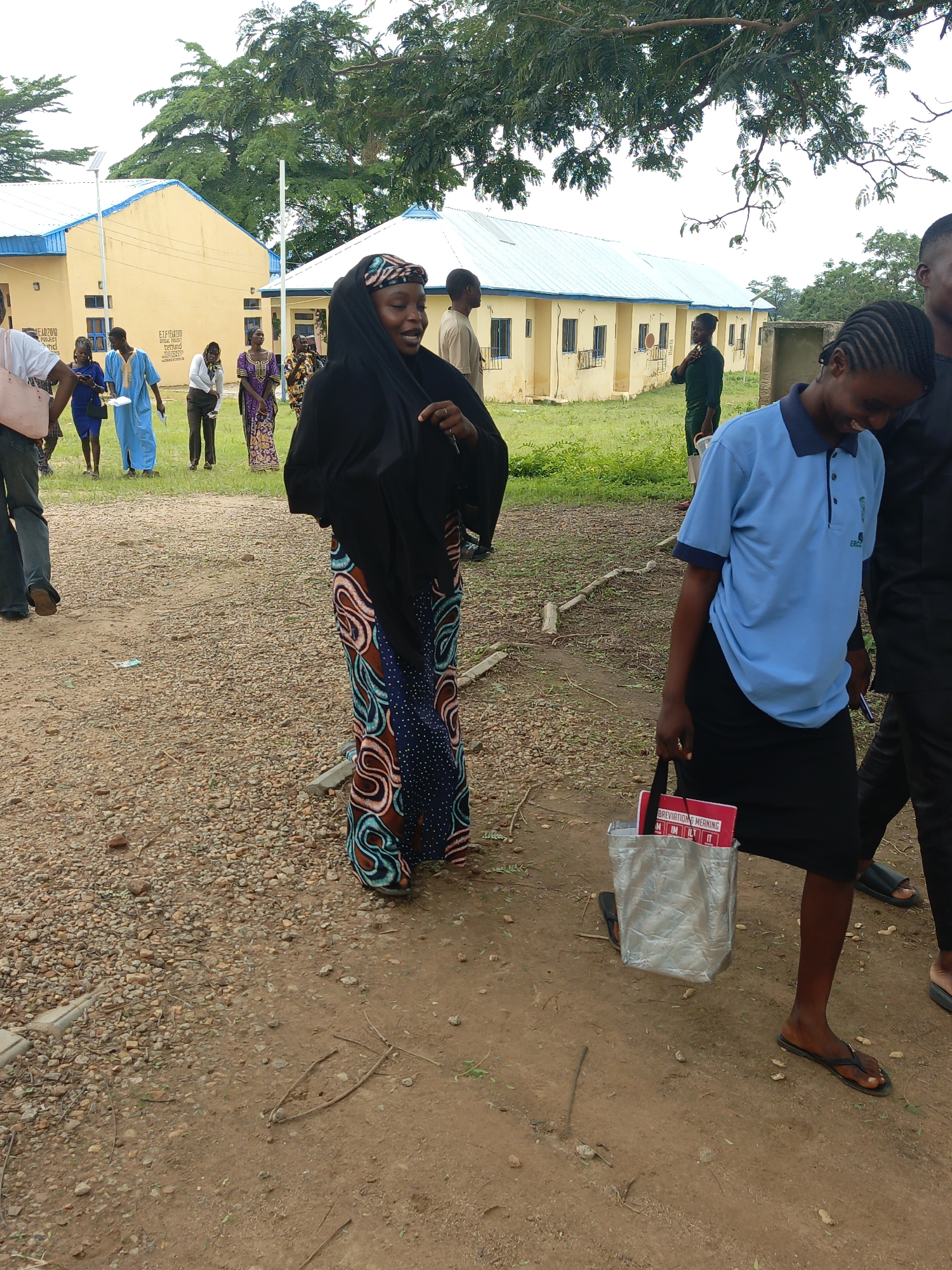 The image shows a group of people walking on a gravel and dirt path in front of a building. The building is yellow with a blue roof and appears to be a school or administrative facility. There are trees and grass in the background.
In the foreground, a woman wearing a black headscarf and a colorful patterned dress is walking towards the viewer, looking to her right. She is holding a key in her hand. To her right, another woman wearing a light blue polo shirt and a black skirt is walking away from the viewer, carrying a silver bag with a red book inside. To the far right, a person in a black shirt and pants is walking away from the viewer.
In the background, several other people are visible, some walking and some standing. They are wearing various types of clothing, including traditional African attire and more casual wear. The overall scene suggests an outdoor gathering or a day of activity at an educational institution.