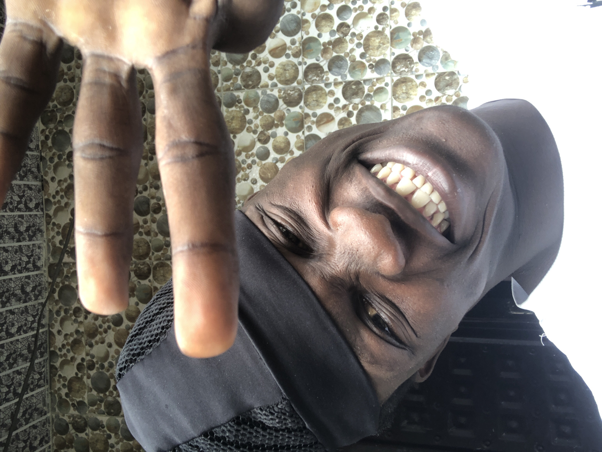 A close-up, slightly tilted selfie shows a Black individual wearing a black durag and a white shirt. The person has dark skin, is smiling widely, and is holding up their right hand, palm facing the camera, with fingers spread. The background features a tiled wall with a pattern of circles in various shades of brown, beige, and grey, and above that, a section of patterned wallpaper. The lighting appears to be natural, coming from the front and slightly to the right, creating highlights on the person's face and hand. The image was taken in Ado, Nigeria.
