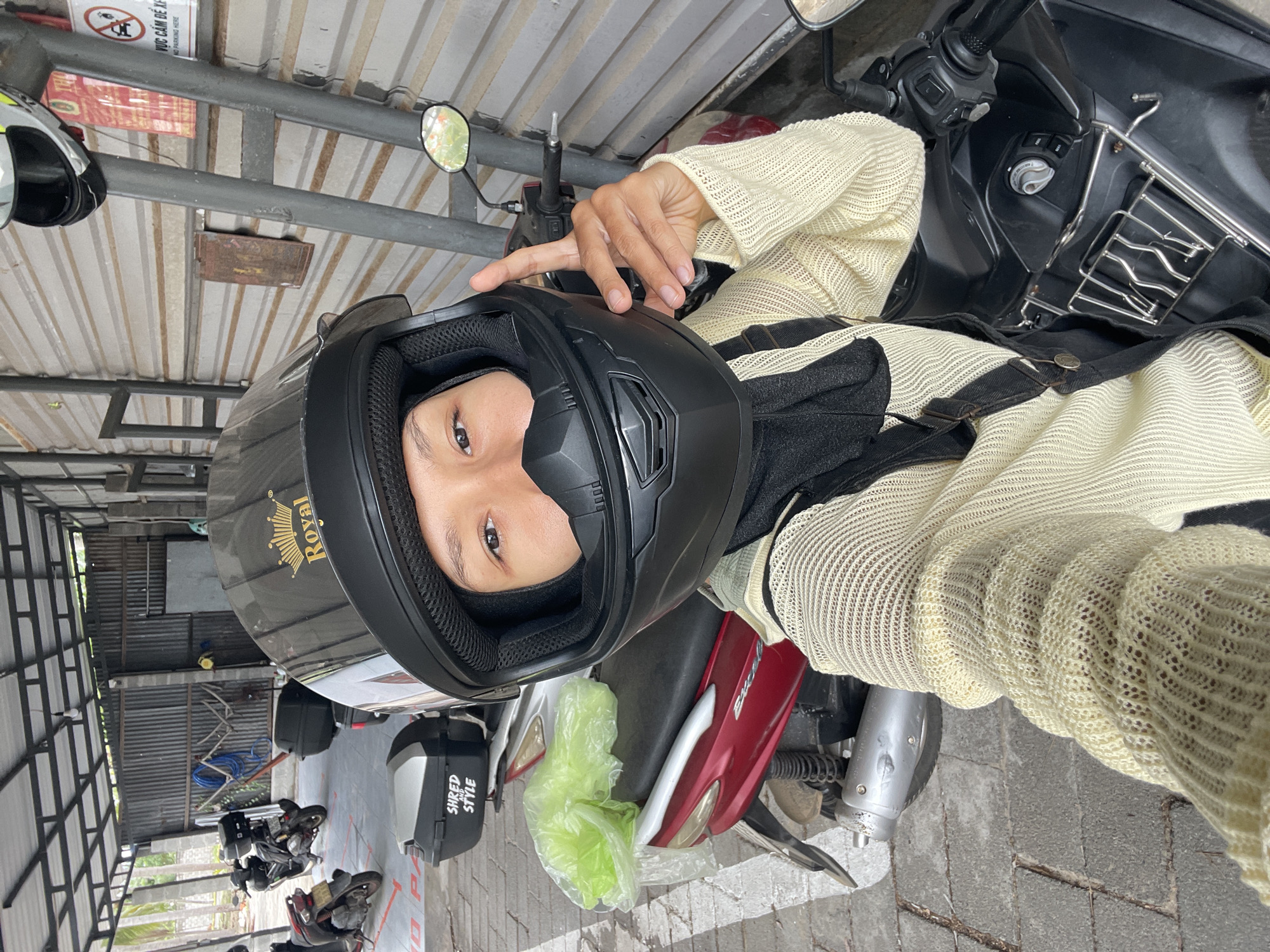 A person wearing a black helmet is seated on a parked motorcycle in a covered outdoor area. The motorcycle is red and black. The person is wearing a light-colored, loosely knit sweater and black overalls. The helmet has the word "Royal" on it. The background shows a corrugated metal roof and other parked motorcycles. In the distance, there are signs with Vietnamese writing. The location is likely in Phu Thuy Ward, Vietnam, based on the language of the signs.