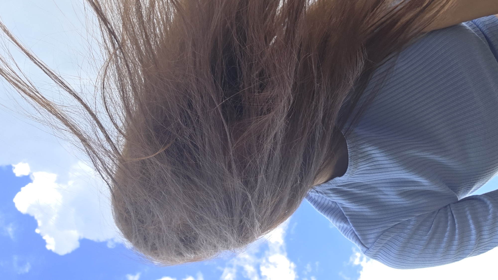 A low-angle, close-up shot captures a person's head and shoulders against a bright blue sky with scattered white clouds. The person's long, light brown hair is blowing across their face and obscuring it. They are wearing a light blue, ribbed, long-sleeved shirt. The scene appears to be outdoors.