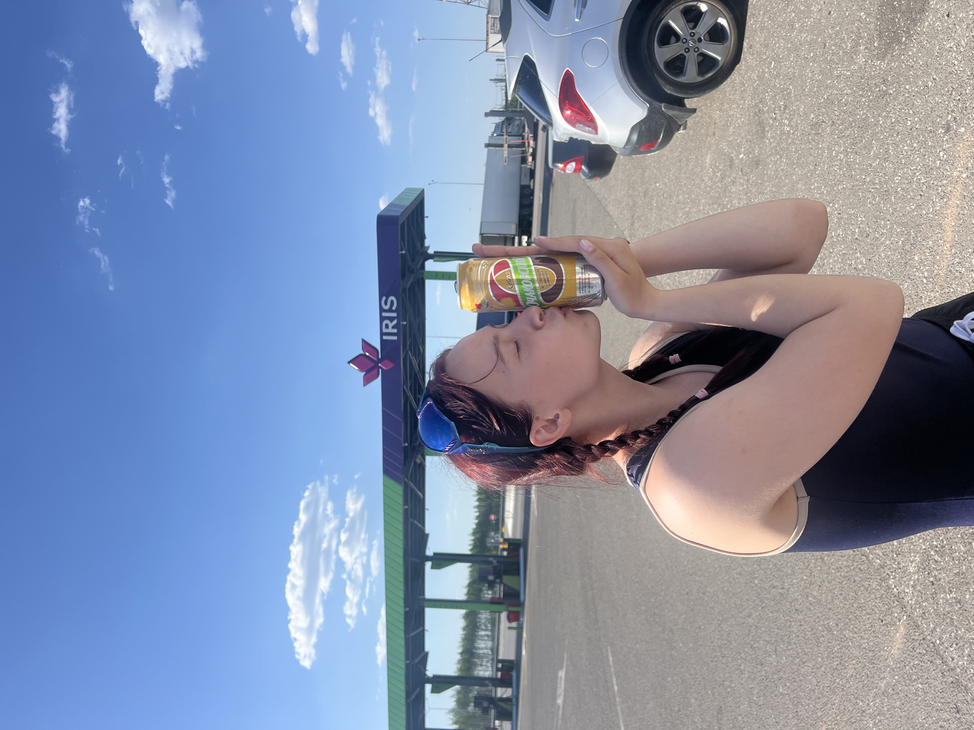 A woman with braided auburn hair and blue sunglasses is standing outdoors, posing with a beverage can near an IRIS toll gate. The location is likely a toll plaza in Казань, Russia, indicated by the "IRIS" sign. The sky is blue with scattered white clouds. A silver car is parked nearby on the asphalt surface. The woman is wearing a dark-colored swimsuit or athletic wear.