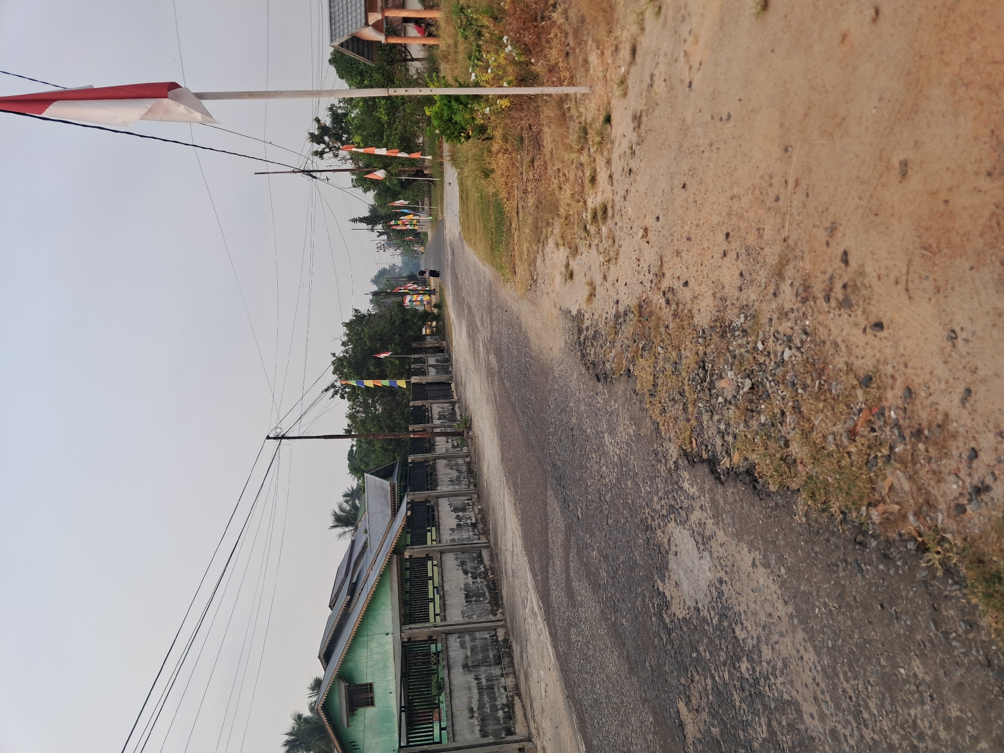The image depicts a street scene in the city of Mempawah, Indonesia, likely during a festive period, given the presence of numerous Indonesian flags. The road, which appears to be asphalt with some wear and tear, curves to the left in the foreground. To the left of the road, a light green building with a dark roof is visible, bordered by a concrete fence with metal railings. Several power lines crisscross the overcast sky.
Along the right side of the road, a dirt and gravel embankment slopes upwards, with dry grass and sparse vegetation. Further down the street, on both sides, are more Indonesian flags, indicating a celebration or commemoration. A person, possibly female due to clothing and headscarf, is walking away from the camera down the street on the left side. In the distance, more buildings and trees can be seen. The overall impression is of a quiet residential street decorated for an event.