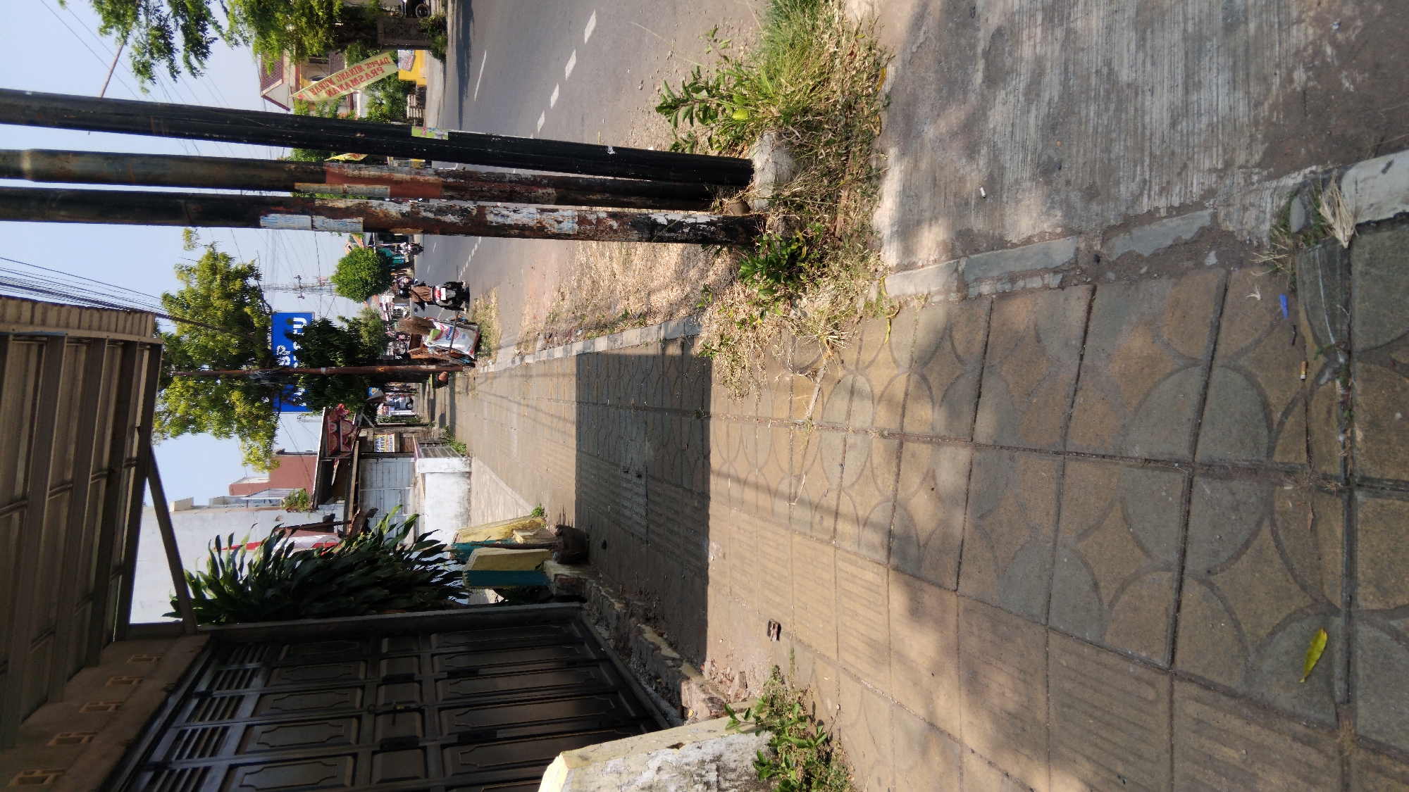 The image depicts a street scene in Proyonangg, Indonesia. In the foreground, a light-colored, textured sidewalk runs alongside a road. The sidewalk is bordered by dry grass and weeds on the right and a dark metal gate and a building on the left. Several utility poles, dark and weathered, stand along the edge of the road. Beyond the sidewalk, a road with white lane markings is visible, with a few motorcycles and their riders present, suggesting traffic. Buildings and trees line the far side of the road. The overall scene is urban and appears to be a typical street on a sunny day.