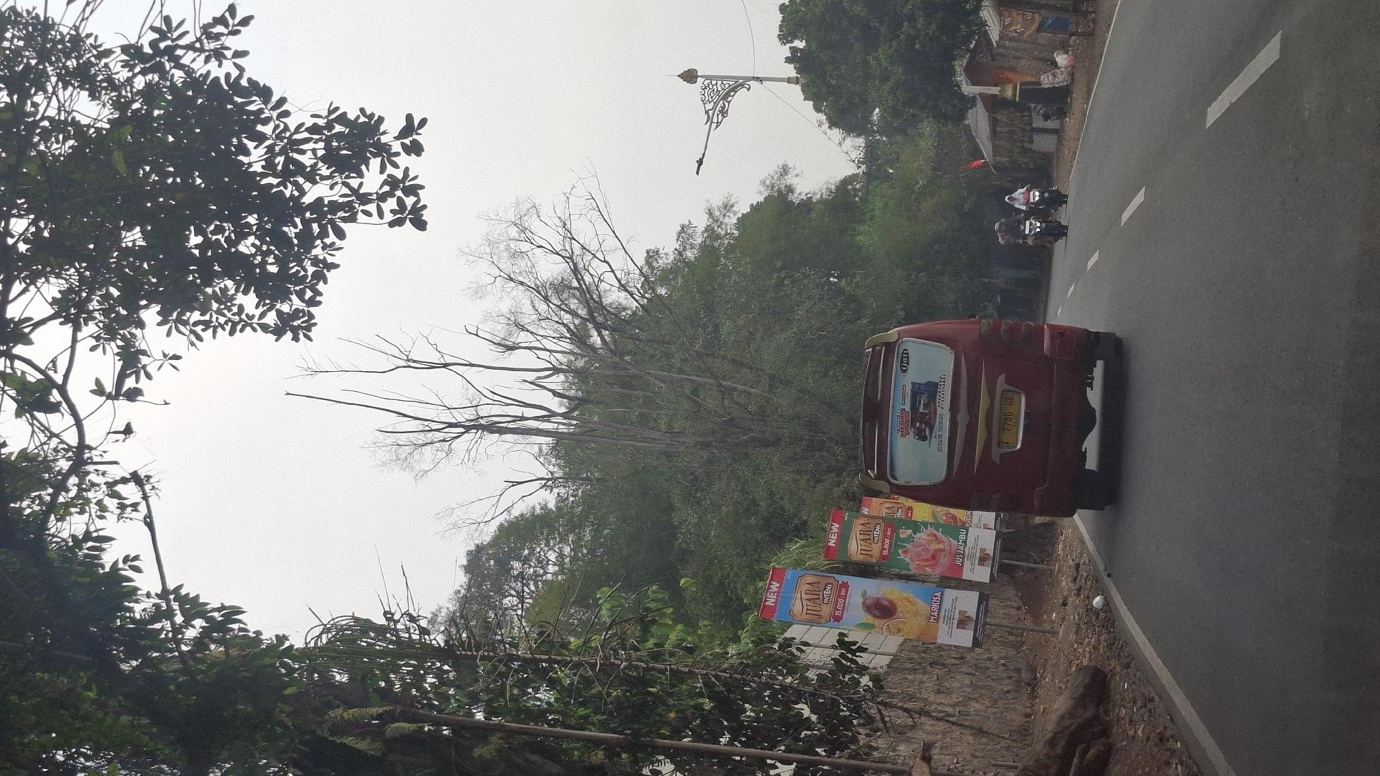 The image captures a road in Cilaku, Indonesia, viewed from a vehicle. On the left side of the road, a stone wall is visible with two large vertical advertisements. The advertisements depict fruit and juice, with the text "JUARA INTENSI" and "MARKISA" and "JUS JAMBU". Further down the road, a red van is driving, its rear window displaying an advertisement for "MERAH PUTIH". Two people on motorcycles are approaching the van from the opposite direction. The environment consists of a paved road, trees and foliage lining the sides, and a cloudy sky. The overall scene depicts a typical day of travel on a road in a semi-urban or rural area of Indonesia.