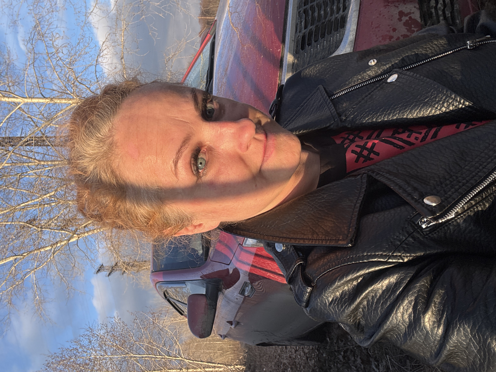 A woman is captured in a close-up selfie, looking directly at the camera with a neutral to slightly smiling expression. Her light-colored eyes (green or blue) are prominent, and her light brown, curly hair appears somewhat disheveled around her face. She is wearing a black leather jacket with silver zippers and snaps, layered over a red shirt. Partially visible black text on the red shirt can be discerned as "#R#".
The scene is illuminated by the warm, low light of either early morning or late afternoon (golden hour), casting soft shadows across her face and highlighting the textures of her skin and jacket.
In the background, the front portion of a red vehicle, possibly a pickup truck or SUV, is visible to her right, appearing somewhat dirty or dusty. Behind the vehicle and extending across the upper left part of the frame are numerous bare tree branches against a clear blue sky with scattered white clouds. The leafless trees suggest the season is late autumn, winter, or early spring. The overall setting appears to be an outdoor, possibly rural or semi-rural environment in Chaikovsky, Russia.