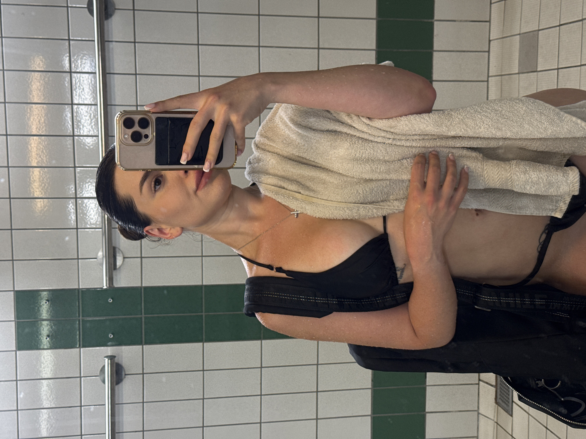 The media file shows a young woman taking a selfie in a tiled room, possibly a locker room or bathroom. She is holding a smartphone in her right hand, angled to capture her reflection. She is wearing a black bikini top and has a light-colored towel draped over her shoulder. Her hair is dark and pulled back. She has a necklace with a small cross pendant. Her skin appears to have water droplets on it, suggesting she may have just showered or been swimming. The walls are covered in white and green tiles, and there are metal fixtures visible, such as a towel rack or shower head. The overall lighting suggests it might be daytime, but it is difficult to determine precisely. The setting appears to be indoors. The woman's expression is neutral. No visible text is present in the image. The location context provided is Prague, Czechia.