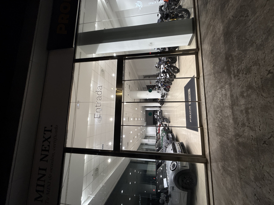 The image depicts the brightly lit exterior of a vehicle showroom at night, located in Polígon Industrial De Son Castello, Spain. The foreground features a dark, textured concrete pavement. The building's facade is composed of large, transparent glass panels framed by dark metallic supports, serving as display windows and a central entrance.
Above the left showroom section, a sign reads "MINI NEXT. Tu MINI de ocasion, certificado" (Your used, certified MINI). Above the main entrance, the word "Entrada" (Entrance) is visible in white lettering on the glass. The entrance consists of automatic sliding glass doors. Immediately inside, a dark rectangular mat bearing "PRO MAX" in white lettering is on the floor. A partial "PRO MAX" sign is also visible on the dark facade above the right section of the showroom.
The interior, well-illuminated by numerous recessed ceiling lights, showcases vehicles on a light-colored tiled floor. The left side displays several Mini Cooper automobiles, including a silver/grey Mini Cooper convertible with black wheels and a visible license plate "6094 MBN." Other Mini Coopers in various colors are arranged behind it. The right side of the showroom displays multiple motorcycles, predominantly dark in color, arranged in rows. Several motorcycles feature golden-colored front forks. No individuals are present, and no actions or interactions are occurring within the scene.