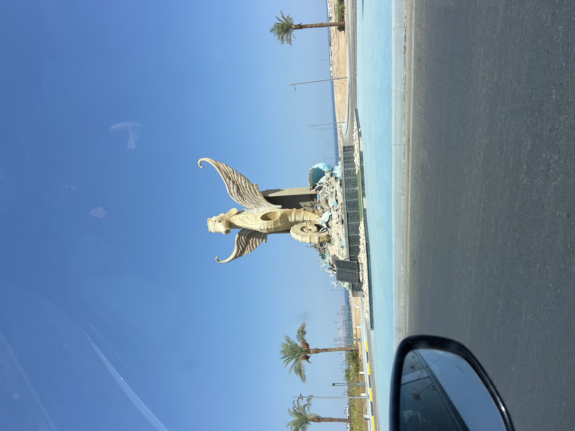 This image captures a monumental sculpture of a winged seahorse, a prominent landmark in Sharm El Sheikh, Egypt. The sculpture, made of a light-colored material, stands on a circular base decorated with rocks and marine life, within a pool of bright blue water. The seahorse sculpture is intricately designed, with detailed wings and a coiled tail. In the background, palm trees line a walkway, and the blue expanse of the sea stretches to the horizon under a clear sky. The scene is viewed from a vehicle, as evidenced by the car's side mirror visible in the lower left corner. The presence of streetlights and paved roads suggests an urban or developed coastal area, characteristic of Sharm El Sheikh's tourist districts.
