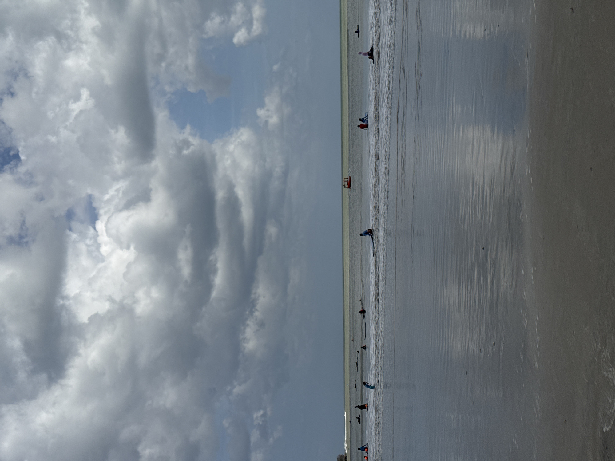 The image depicts a crowded surf beach in Kuta Selatan, Indonesia, on a cloudy day. The foreground features wet sand with ripples and scattered debris, transitioning into shallow water with gentle waves. In the middle ground, numerous individuals are scattered across the water, many on surfboards, with some paddling and others sitting or preparing to surf. A small orange boat is visible further out in the ocean. The background is dominated by a vast expanse of ocean under a heavily clouded sky, with a sliver of brighter sky visible towards the right. The overall scene is one of recreational activity at the beach.