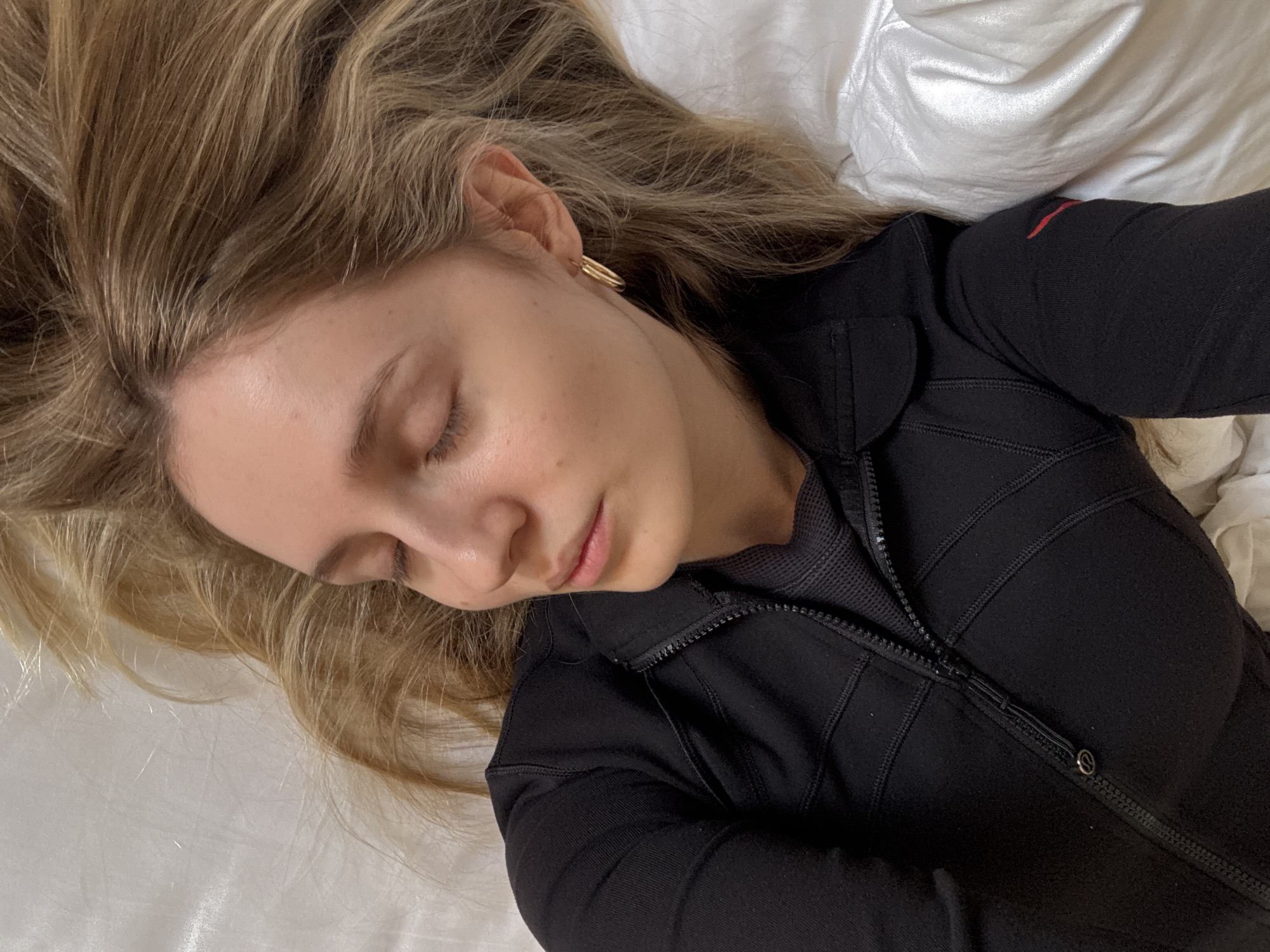 A single individual with light skin, closed eyes, and long blonde hair is depicted lying on their back on a white bed with a white pillow. The person wears a black, long-sleeved athletic top with a full-front zipper, partially unzipped to reveal a black mesh lining. The zipper pull features a small, circular emblem. A subtle red detail is visible on the left upper arm of the top. A single gold-colored hoop earring is present on the right earlobe. The scene is indoors, lacking specific identifying features to determine a precise location within Mexico City, Mexico.