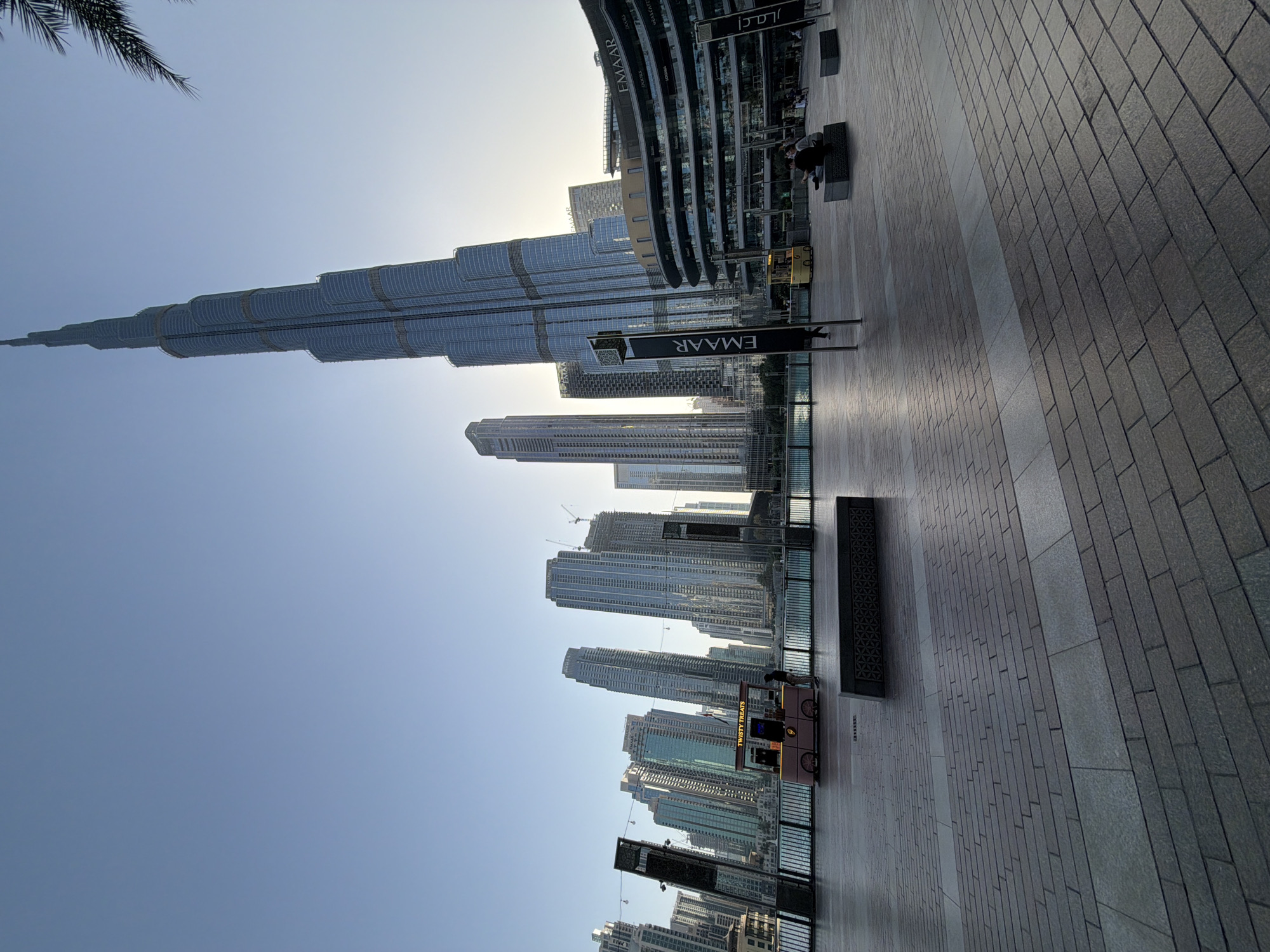 This photograph depicts the Burj Khalifa, a skyscraper in Dubai, United Arab Emirates. In the foreground is a pedestrian walkway paved with gray bricks. Various modern buildings stand behind the walkway. A food cart is located on the walkway. An "EMAAR" sign is visible as well. The sky is blue and clear.