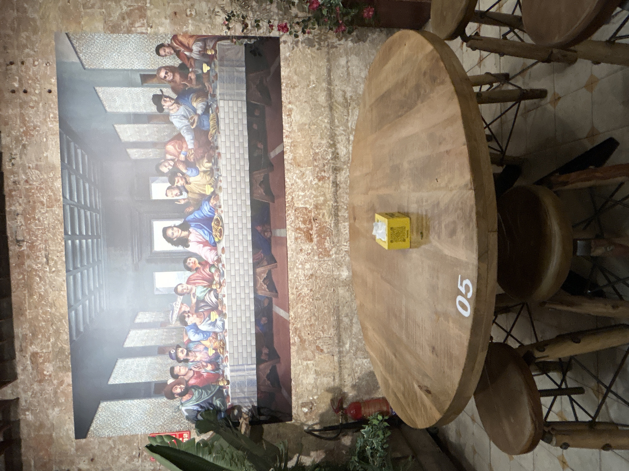 The image depicts an interior scene of a restaurant or bar. Dominating the background is a large mural replicating Leonardo da Vinci's "The Last Supper," featuring a stylized group of people seated at a long table. The mural is set against a brick wall. In the foreground, a round wooden table is centrally placed, with several wooden stools surrounding it. A yellow box of tissues rests on the table. The floor appears to be tiled. To the left, a fire extinguisher and a potted plant are visible. The overall ambiance suggests a casual dining or gathering space. The location in Barcelona, Spain is not discernible from the provided image.
