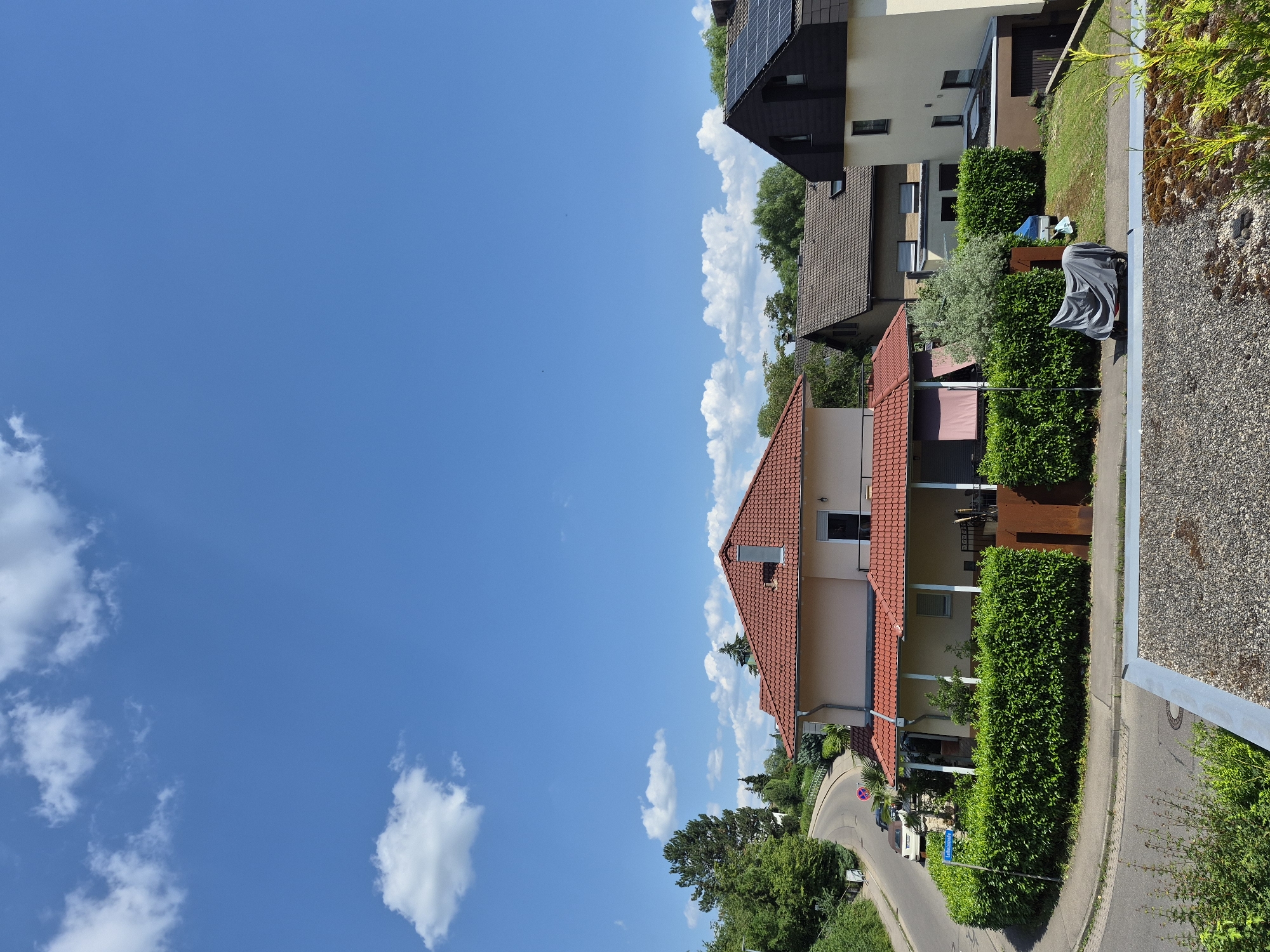 The image shows a residential street scene on a sunny day with a clear blue sky and scattered white clouds. In the foreground, a grey tarp covers a wheeled object, possibly a motorcycle or a scooter, parked on a paved area. Behind this, a well-maintained, dense green hedge runs along the edge of a lawn. Several houses are visible, varying in architectural style and color.
The house on the left has a prominent red tiled roof and a light-colored facade. It features a covered porch area with white pillars. A street sign reading "Lautsbergstraße" is visible on the left side of the road. A curved road with some parked cars winds its way up a green, tree-lined hill to the left of the houses.
Further back and to the right, another house with a brown tiled roof and dormer windows is partially visible. To its right is a modern-looking house with a dark facade and solar panels on its roof. This house has several windows and a garage entrance. The overall environment suggests a suburban or semi-rural setting. The scene is tranquil, with no people or animals visible. The time of day appears to be midday, given the bright sunlight and clear sky.