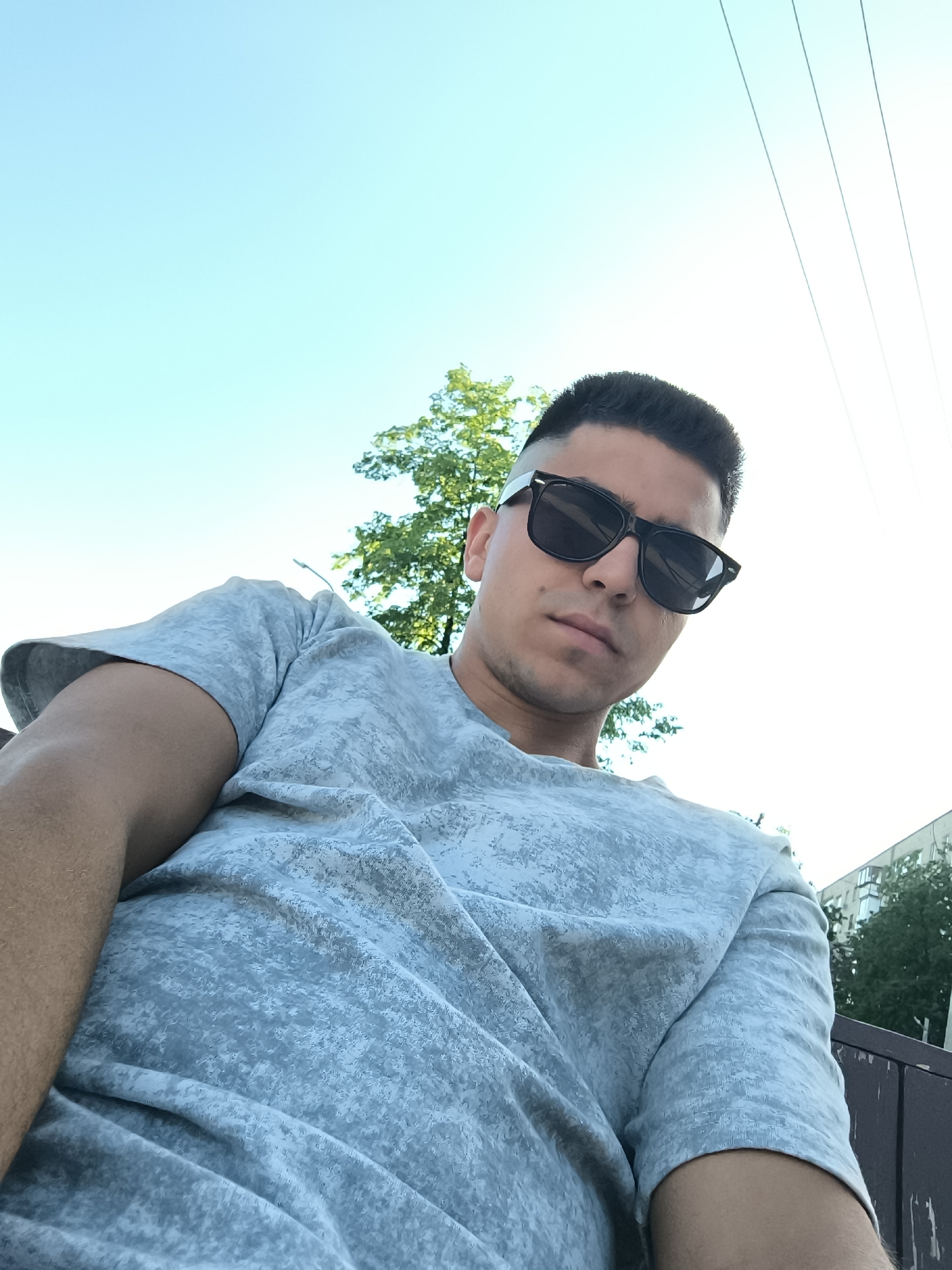 A low-angle, full shot shows a man wearing dark sunglasses and a gray, short-sleeved t-shirt. He looks directly at the camera. Behind him, there is a tree and a clear sky. Power lines are visible in the upper right corner. A building is partially visible on the right side of the frame. Part of a dark brown structure, likely a bench or fence, is in the lower right. The specific location within the city of Unknown, Unknown cannot be determined with the available information.