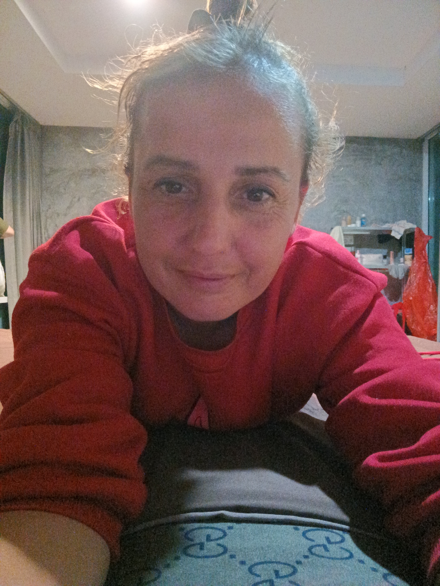 A medium shot of a woman wearing a red sweatshirt, lying on a bed with a blue and white Gucci-patterned pillow. She faces the camera with a slight smile. Her hair is up, some strands sticking out. The background shows a gray concrete wall, a curtain on the left, and a shelf with various objects including a red plastic bag on the right. The location is implied to be in Ban Tak Daet, Thailand.