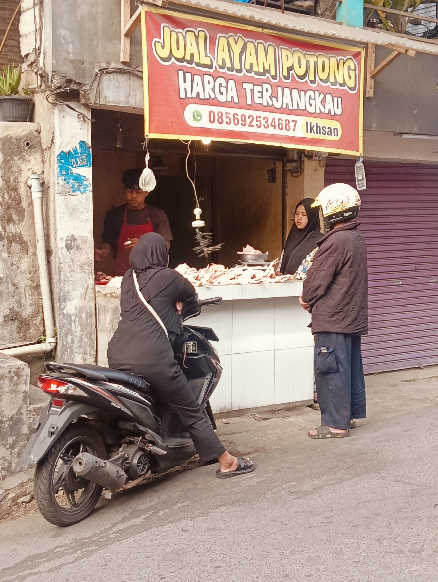 In an outdoor market setting in an unknown city, a butcher stall is shown with a large red banner overhead advertising "JUAL AYAM POTONG HARGA TERJANGKAU" (Chicken Selling, Affordable Price) with a phone number and name. A young man in a red apron stands behind the counter, attending to raw chickens displayed on a white surface. A woman wearing a black hijab sits on a black motorcycle parked in front of the stall, facing away from the viewer. To the right of the stall, a man wearing a helmet and a dark jacket stands facing the stall, appearing to interact with a woman who is partly visible behind the counter, also wearing a black hijab. The environment includes a weathered building facade to the left and a purple shuttered storefront to the right. The ground is paved with asphalt.