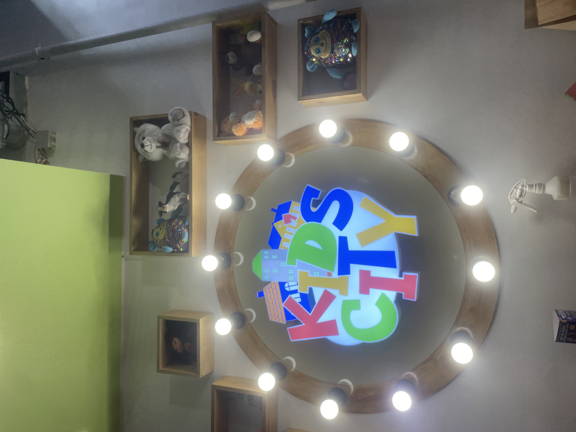 A sign that reads "KIDS CITY" in large, colorful block letters is displayed prominently on a wall. The letters are outlined in white and filled with vibrant colors such as pink, yellow, green, and blue. Behind the letters, a stylized image of a town with colorful houses and rooftops is visible. The sign is illuminated by a circular arrangement of light bulbs around its perimeter. Above and to the sides of the sign, several wooden shelves are mounted on the wall. These shelves hold various stuffed animals, including a white bear, a black and white cow, a fluffy blue creature, and other assorted plush toys. The wall itself is painted a neutral, light gray color, and the ceiling above is painted a bright, lime green. The overall setting appears to be an indoor space, possibly a children's entertainment center or a store, located in Vladivostok, Russia.