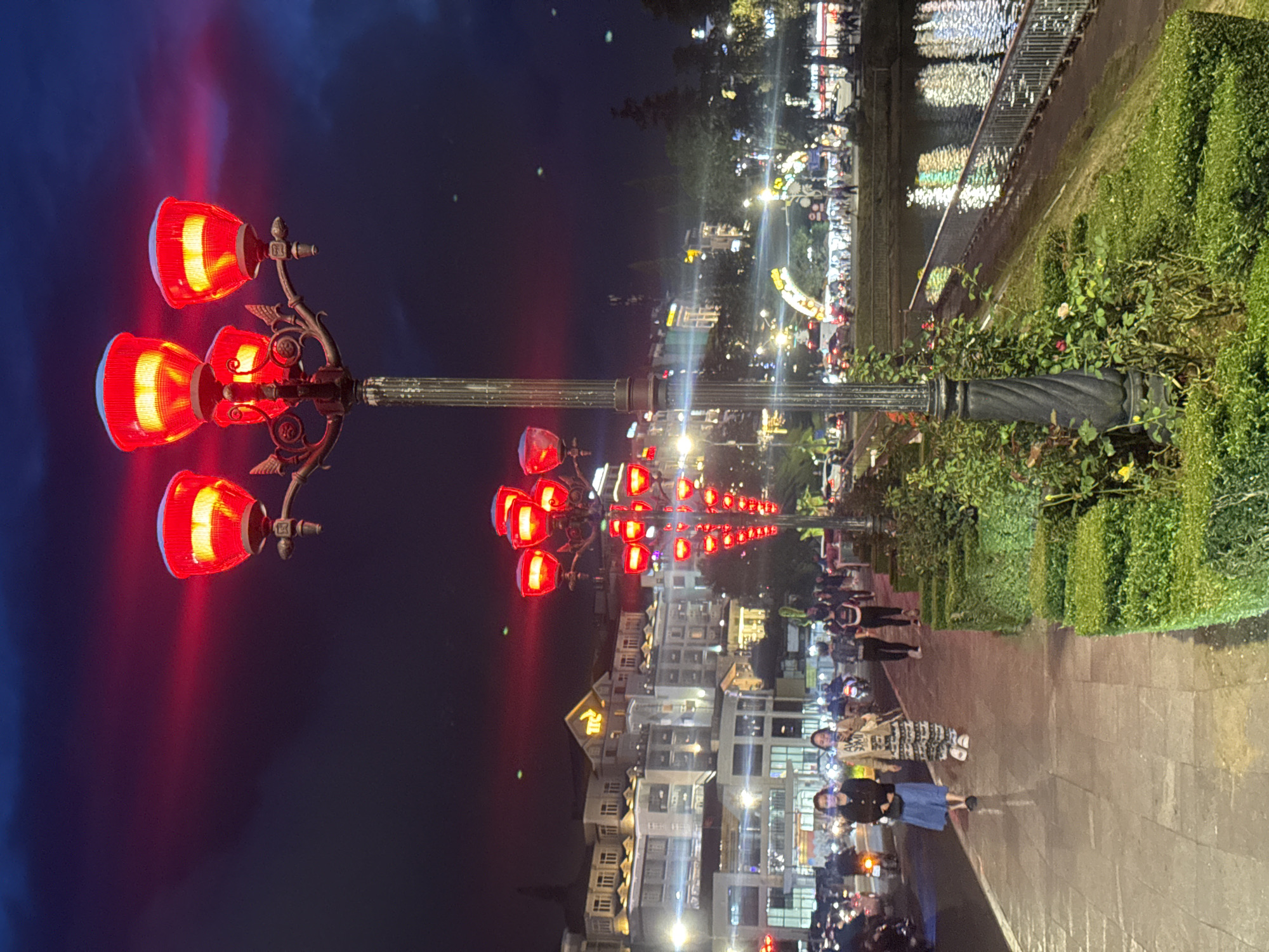 The image shows a street scene at night in Da Lat, Vietnam. A row of red-lit decorative lampposts stands along a walkway. Buildings are visible in the background, illuminated by white lights. Pedestrians are present on the walkway, including some wearing face masks. There is a body of water on the right side of the image, with reflective lights along its edge. Greenery is also visible along the walkway.