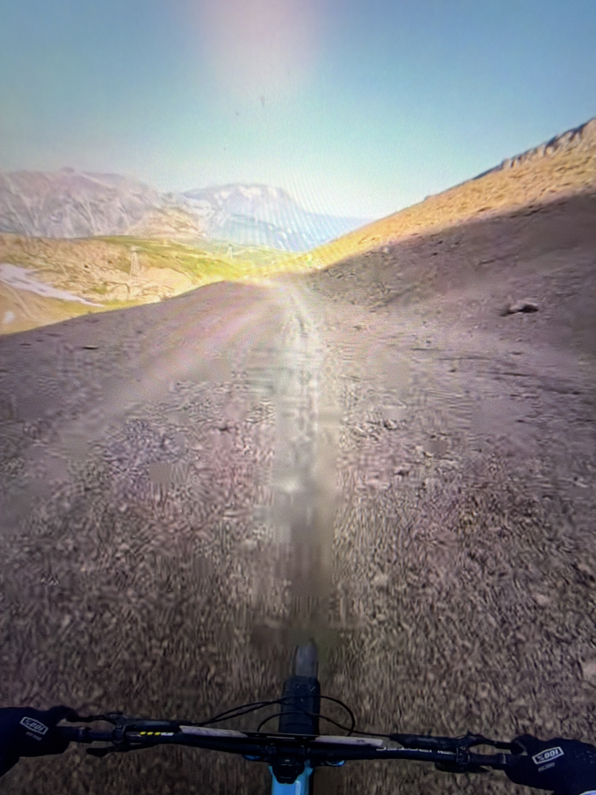 The image is a first-person perspective from a mountain bike, looking down a dusty, rocky trail in a mountainous, arid environment. The sky is a pale blue, with the sun casting a bright glow and lens flare, suggesting it is daytime. The trail appears to be downhill, with loose gravel and dirt. In the distance, there are mountains with sparse vegetation. The handlebars of the bicycle, along with gloved hands gripping them, are visible at the bottom of the frame. The scene depicts an outdoor recreational activity. There is no indication of the city of Kafr Yasif, Israel, in the image.