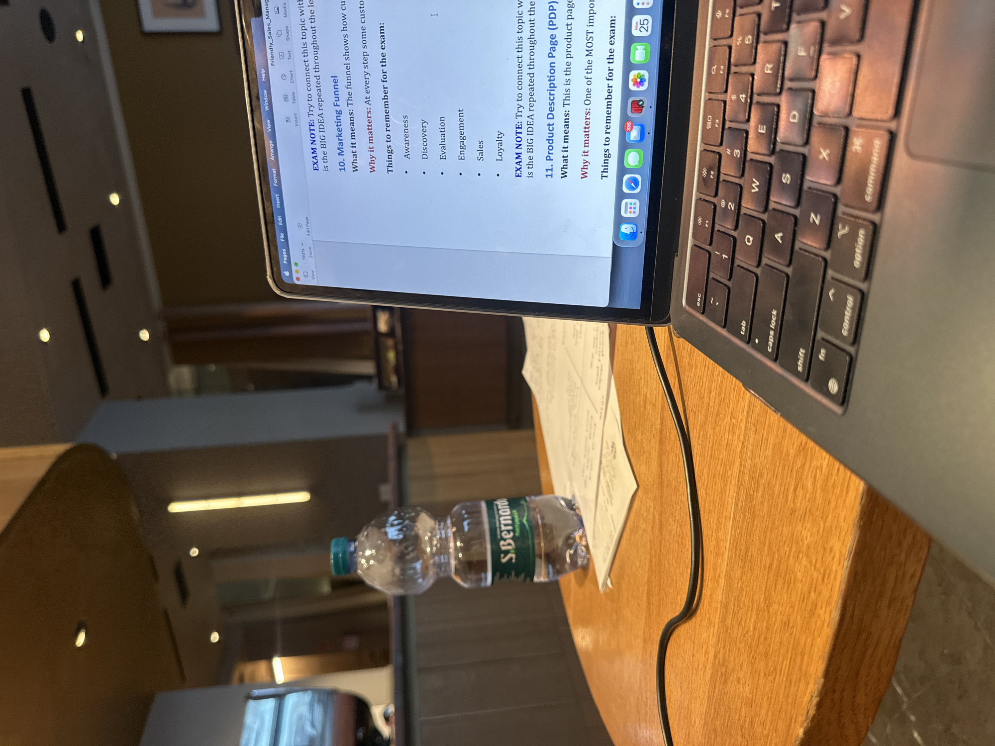 The image shows a laptop sitting on a wooden table, with a bottle of "S.Bernardo" brand mineral water nearby. The laptop screen displays notes about marketing concepts, specifically "Marketing Funnel" and "Product Description Page (PDP)". The laptop keyboard is visible. Background details suggest an indoor location, potentially a cafe or study area, with neutral-toned furnishings and warm lighting. Given the bottle's brand, it is most likely located in Italy, and based on the context of a cafe or study area, the location is very likely Milan, Italy.