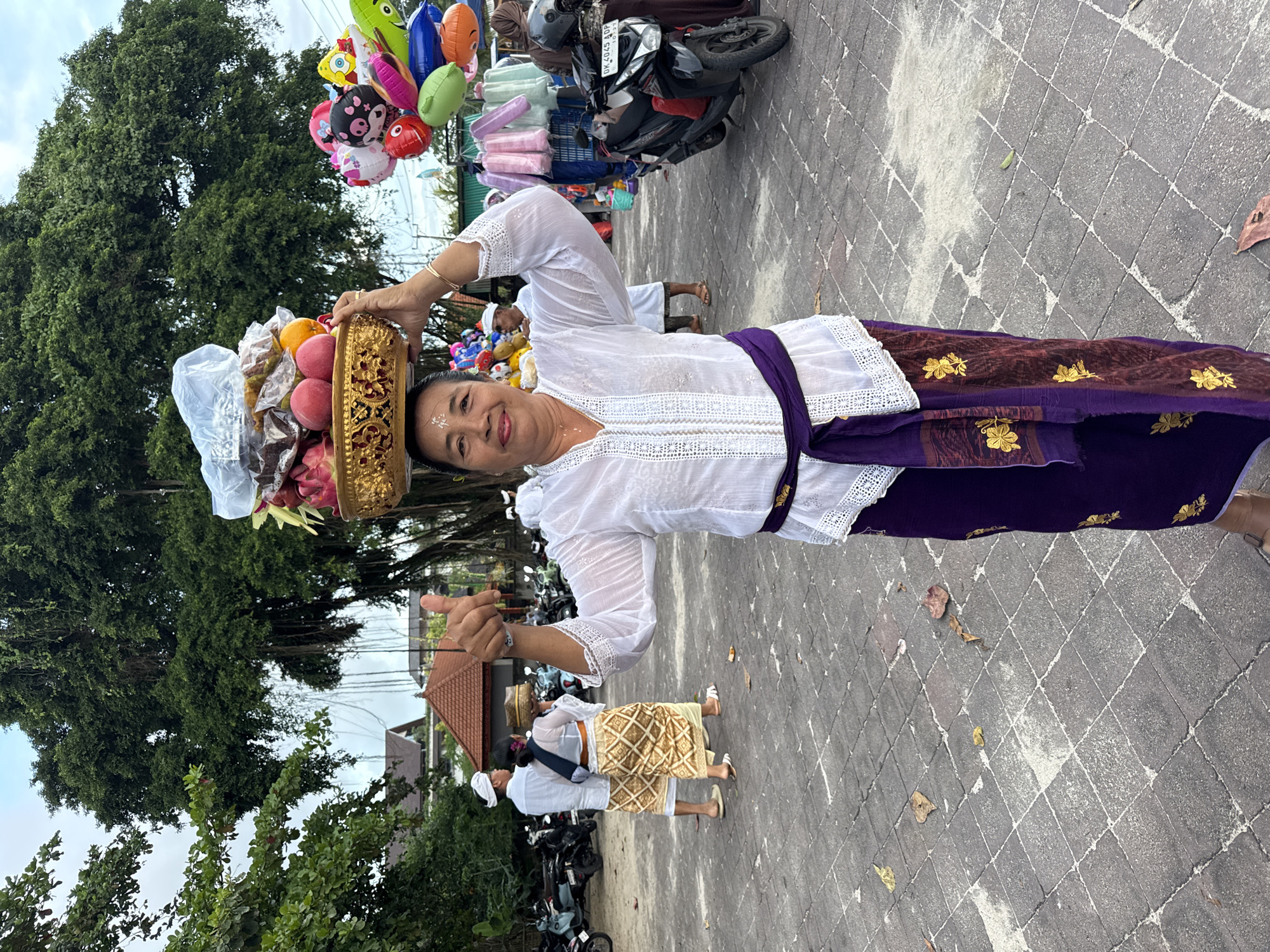 The image captures a vibrant outdoor scene in Kuta, Indonesia, featuring a smiling woman in traditional Balinese attire. She stands prominently, giving a thumbs-up gesture with her left hand, while gracefully balancing an ornate golden basket on her head. The basket is filled with colorful fruits, including peaches, oranges, and dragon fruit, along with other items possibly wrapped in clear plastic, likely offerings.
The woman wears a white, long-sleeved lace kebaya top, a deep purple sarong adorned with intricate gold floral patterns, and a matching purple sash (selendang). A white marking is visible on her forehead, a common feature in Balinese spiritual practices.
The ground is paved with grey interlocking tiles. In the background, large, lush green trees dominate the upper portion of the frame, indicating a natural setting. Parts of a building with a red-tiled roof are visible further back. Other individuals, also dressed in traditional Balinese white tops and patterned sarongs, can be seen in the mid-ground. Several motorcycles or scooters are parked or present; one has a partially visible license plate reading "DK 4045 ADP," confirming a Bali location. To the upper left, a vendor displays an array of colorful balloons featuring various shapes and characters, adding a festive touch to the scene.
The natural lighting suggests it is daytime, under a partly cloudy sky. The woman's cheerful expression and gesture convey a friendly and inviting demeanor, capturing a moment of cultural daily life in Bali.