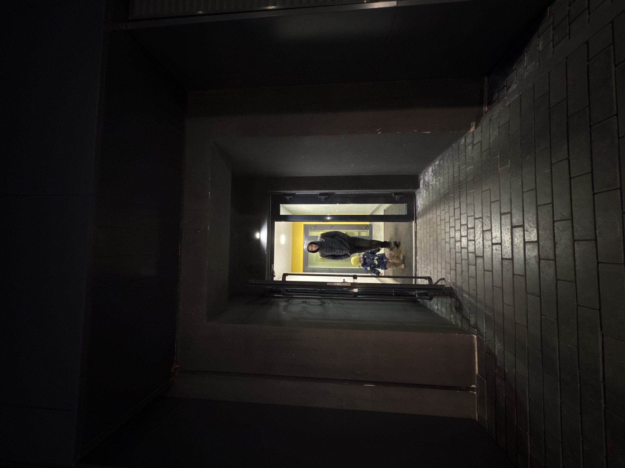 The image shows a dimly lit entryway or tunnel leading to an illuminated interior. The tunnel is constructed from dark-colored stone or brick, with a brick pattern visible on the right wall. The left side of the tunnel is a darker, possibly painted surface. At the end of the tunnel is a doorway or entrance with a brightly lit yellow interior visible. Standing in the doorway are an adult and a child, both dressed in winter clothing. The child wears a yellow hat. The location is possibly Izhevsk, Russia, based on details visible in the original prompt that are not in the picture.