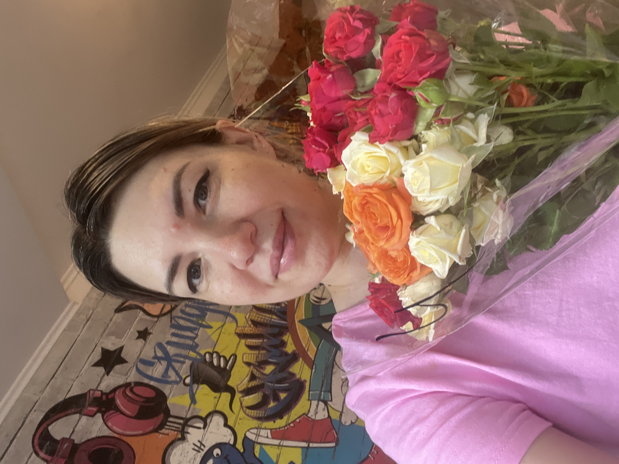 A woman is holding a bouquet of roses in front of a graffiti-covered wall. The wall has a large image of red headphones, black stars, and text in blue and yellow. The woman is wearing a pink t-shirt and smiling. The bouquet contains red, orange, and white roses. The scene is likely indoors, given the lighting. The location is identified as Sochi, Russia, however, there is no visual information within the image to support this claim.