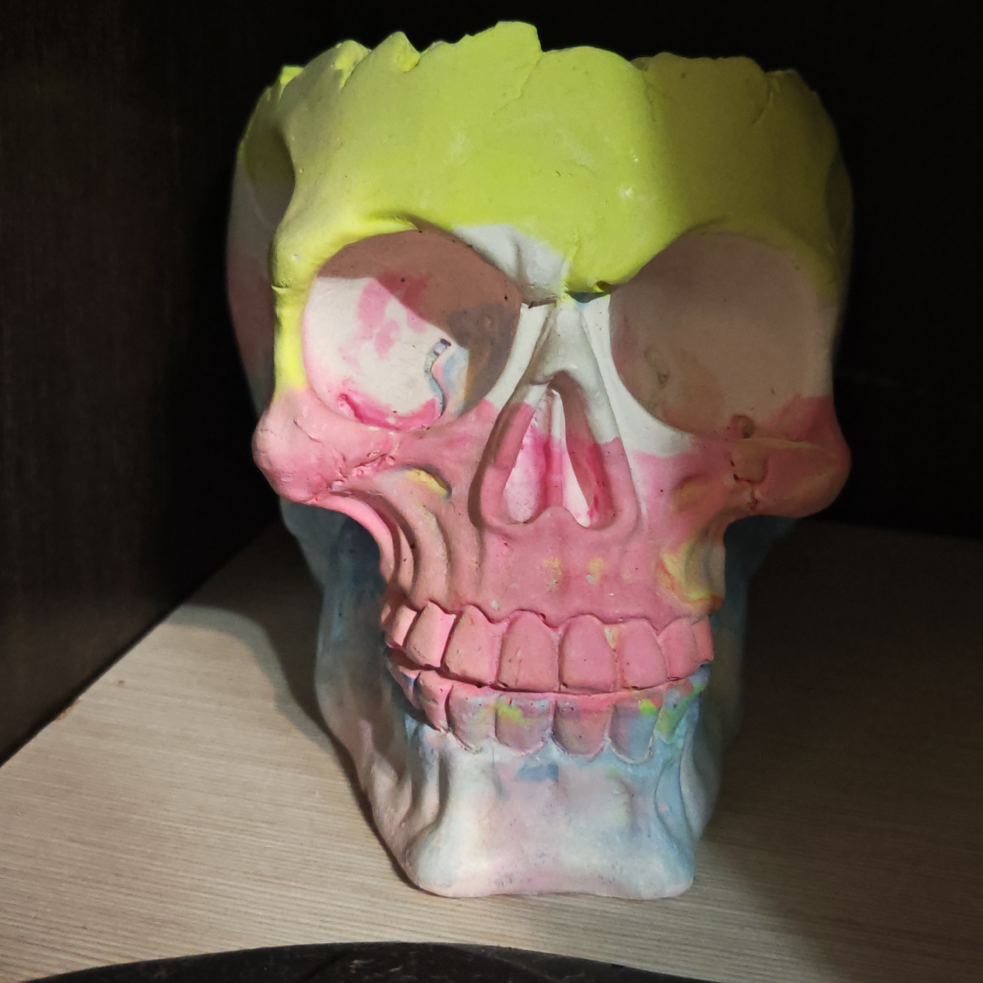 An eye-level shot captures a colorful skull-shaped decorative object, likely a planter or sculpture, standing on a light-colored wooden surface. The skull is composed of various vibrant, pastel hues, applied with a somewhat rough, textured finish. The top portion, resembling the cranium, is predominantly a bright, neon yellow or lime green. Below this, the eye sockets are a light pink and white, with hints of blue. The mid-face, including the cheekbones and nasal cavity, is primarily pink, fading into lighter pinks and some white. The teeth are also pink, with subtle yellow and blue details, indicating an uneven painting style. The lower jaw and chin transition into light blue and white tones.
The object is positioned slightly off-center to the right, facing directly towards the viewer. It casts a soft shadow on the wooden surface behind and to its left. The background is dark and out of focus, suggesting a shadowed corner or the interior of a shelf or cabinet. There is no discernible activity taking place, as it's a still-life image of an object. No visible text is present. The general setting suggests an indoor environment in Novo-Nikol'skoe, Russia. The lighting is focused on the front of the skull, highlighting its bright colors against the dark surroundings.