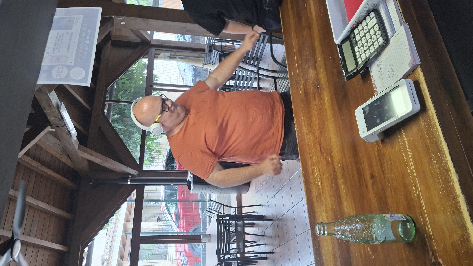 A bald, middle-aged man wearing a bright orange t-shirt, dark shorts, glasses, and white headphones is dancing indoors. He is standing behind a wooden counter. On the counter, there is an empty glass bottle, a credit card reader, a calculator, and a small notepad with writing on it. In the background, there are tables and chairs, a red car parked outside, and large windows. The scene appears to be a casual indoor dining or bar area. The location is the city of La Fresneda, Spain.