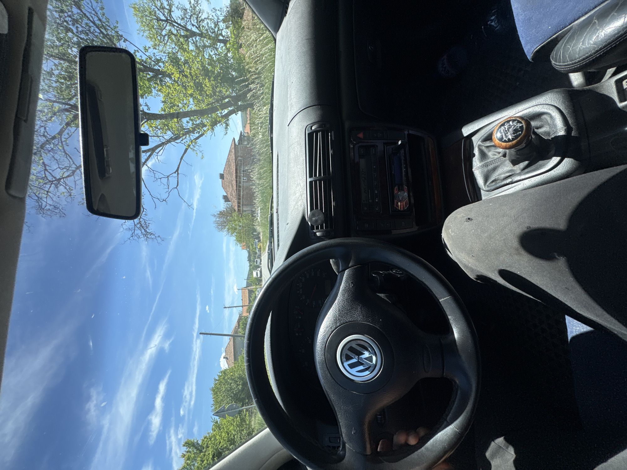 The image shows the interior of a car, specifically the driver's side. The steering wheel, dashboard, rearview mirror, and center console are visible. The steering wheel has a Volkswagen logo. The gear shift has a wooden knob with an emblem on top. Outside the windshield, a residential area with trees and a house can be seen. The sky is blue with white clouds. The image was taken in Batanovtsi, Bulgaria.